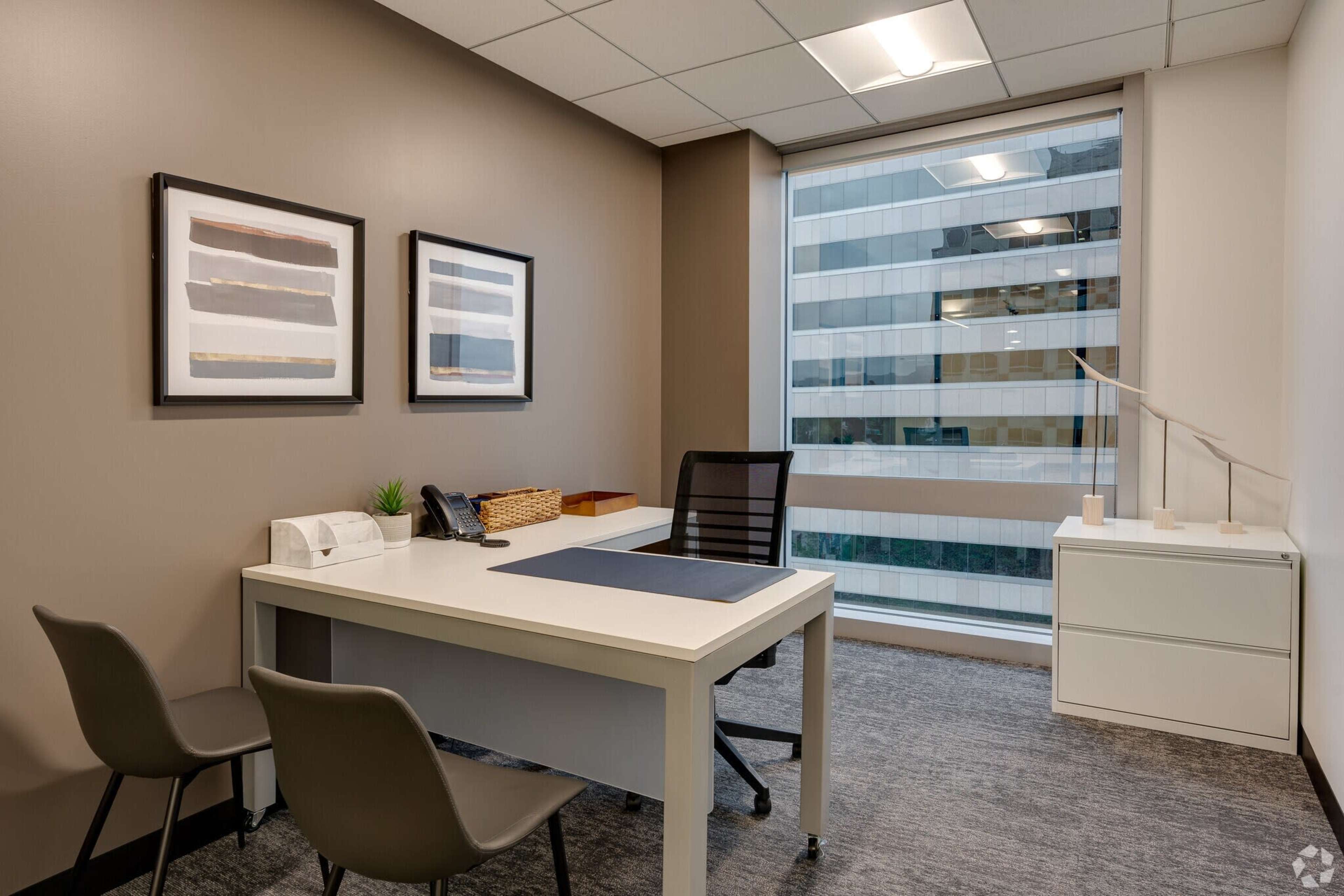 An office space featuring a desk with a chair, a telephone, and framed artwork on the walls, with a window revealing a view of a cityscape.