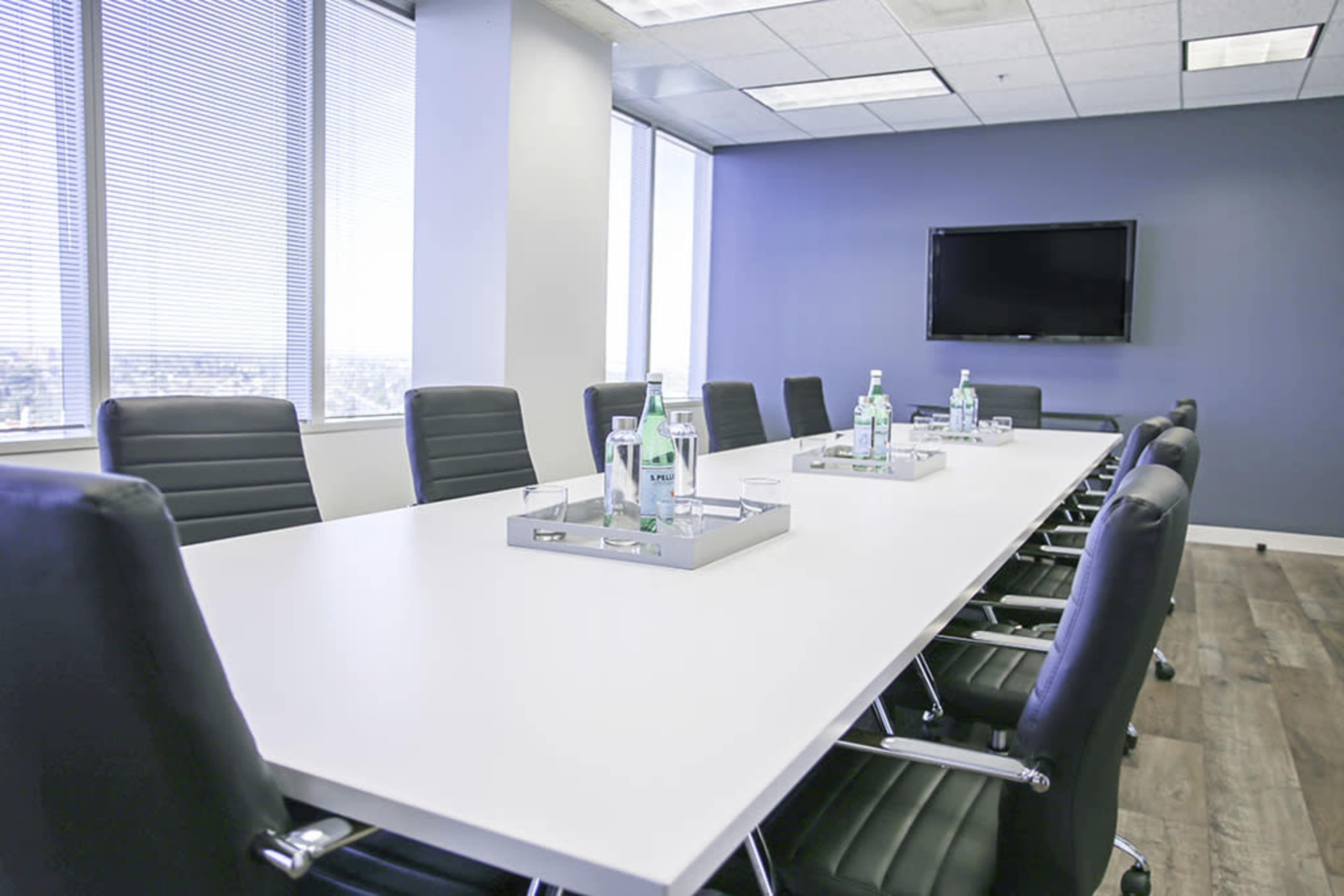 A conference room features a long white table with black chairs, surrounded by windows and a television mounted on a blue wall.