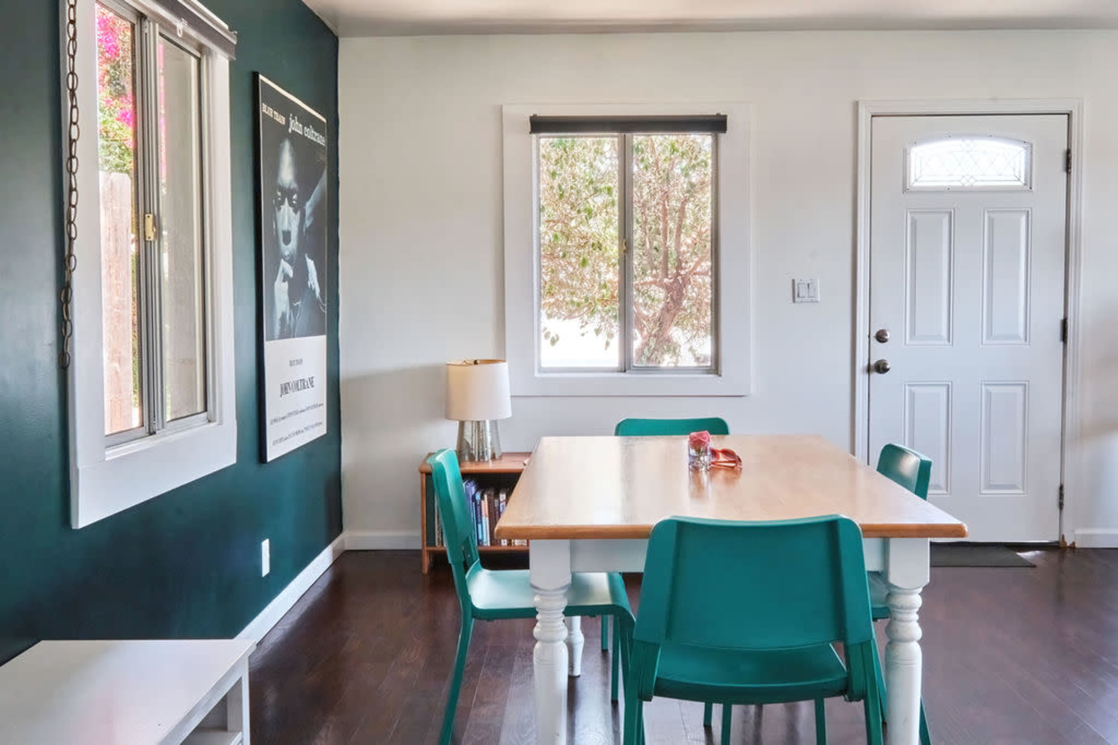 A dining area features a wooden table surrounded by turquoise chairs, with a poster on the wall and a door leading outside.