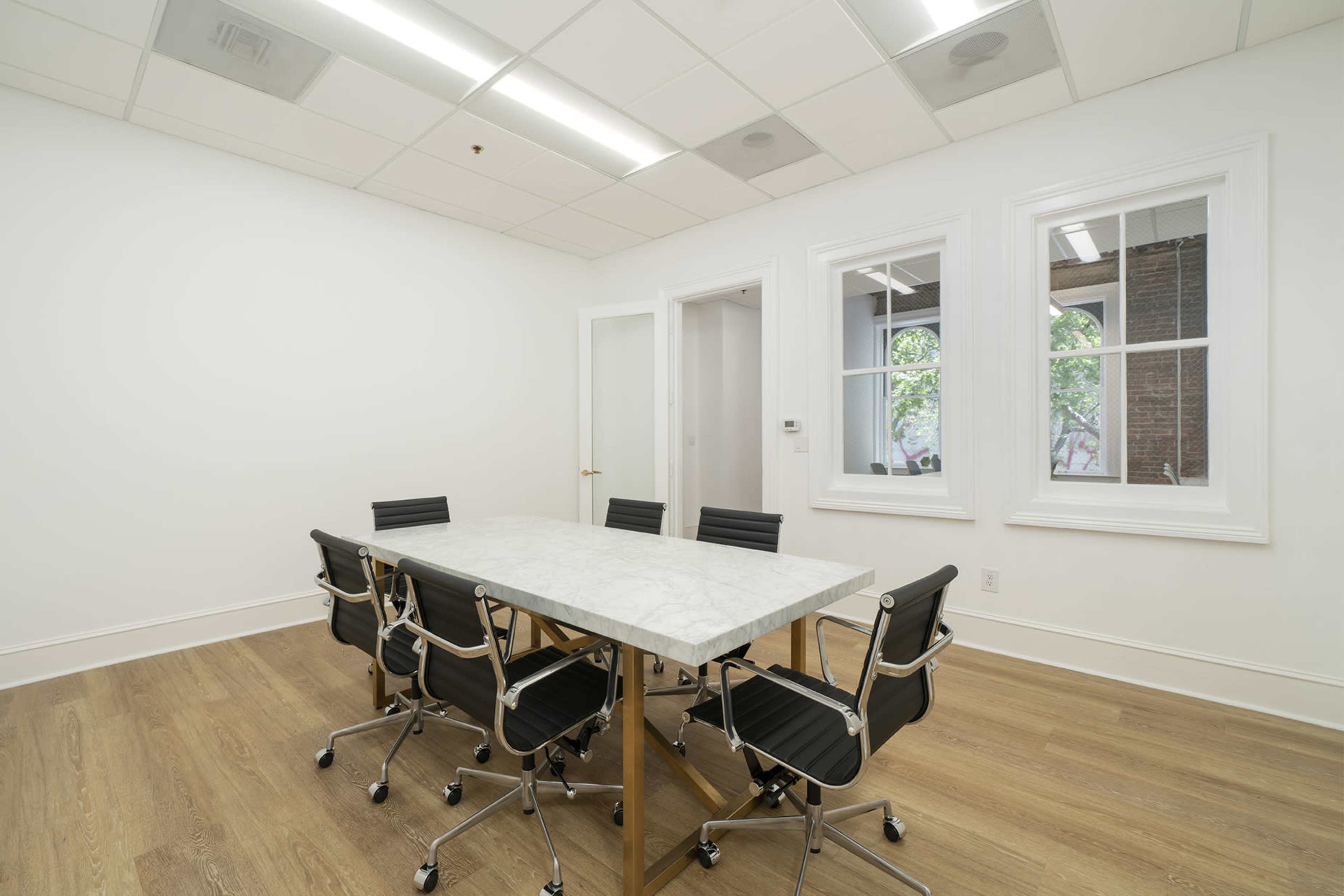 A conference room features a marble-topped table surrounded by six black chairs, with two windows illuminating the space.