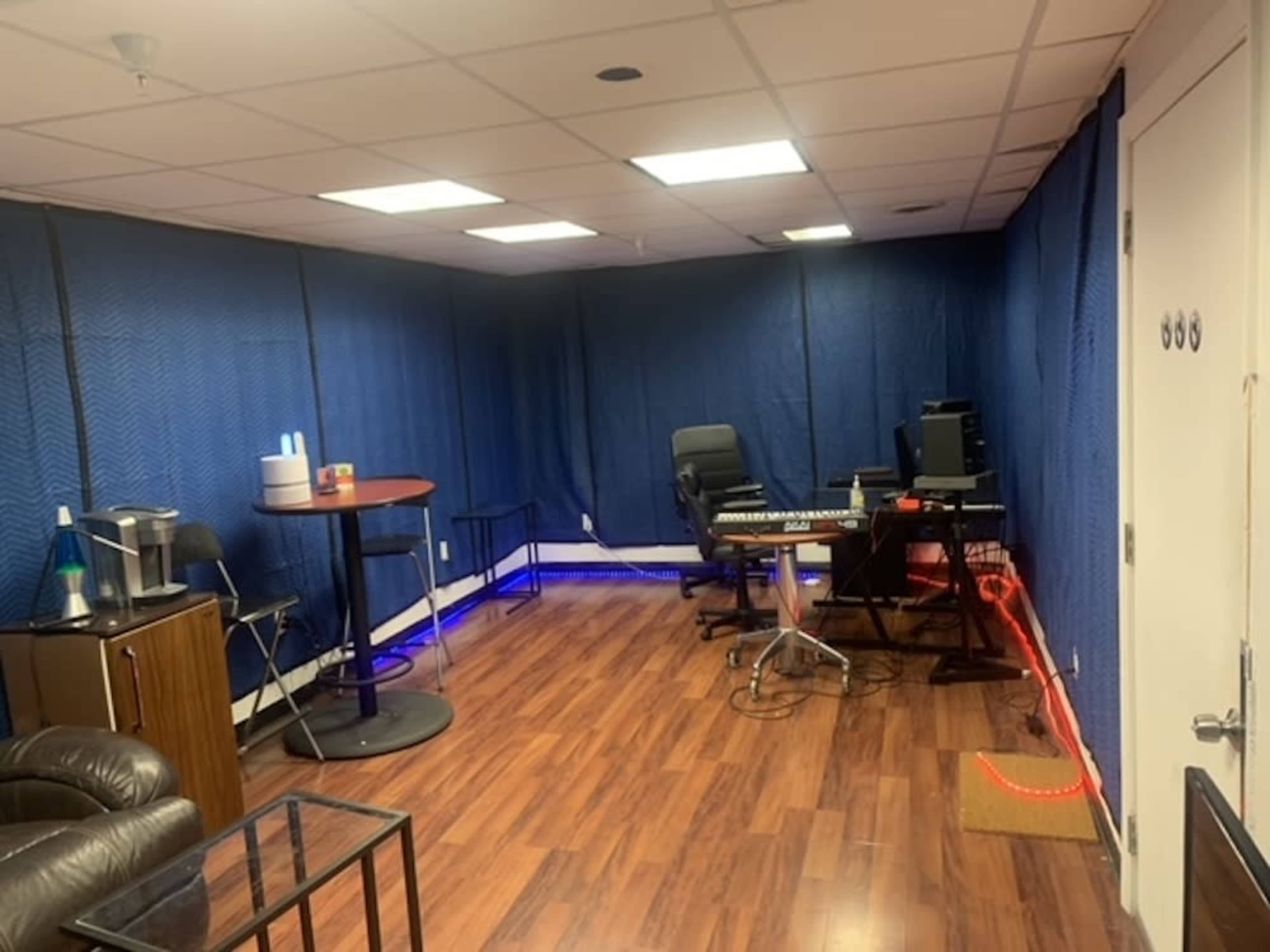 A sparsely furnished room with blue curtains, a black chair, a keyboard setup, and a small table near a coffee station.