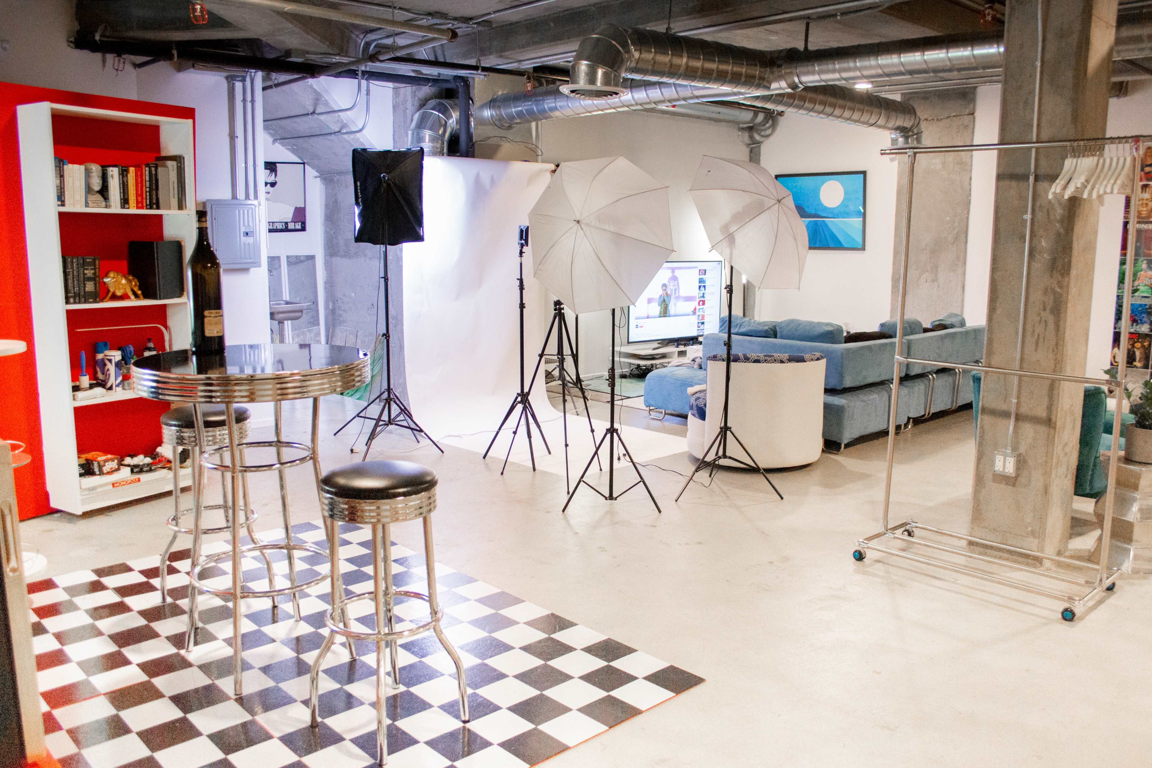 A photography studio setup featuring a checkered floor, two silver bar stools, softbox lights, and a couch in the background.