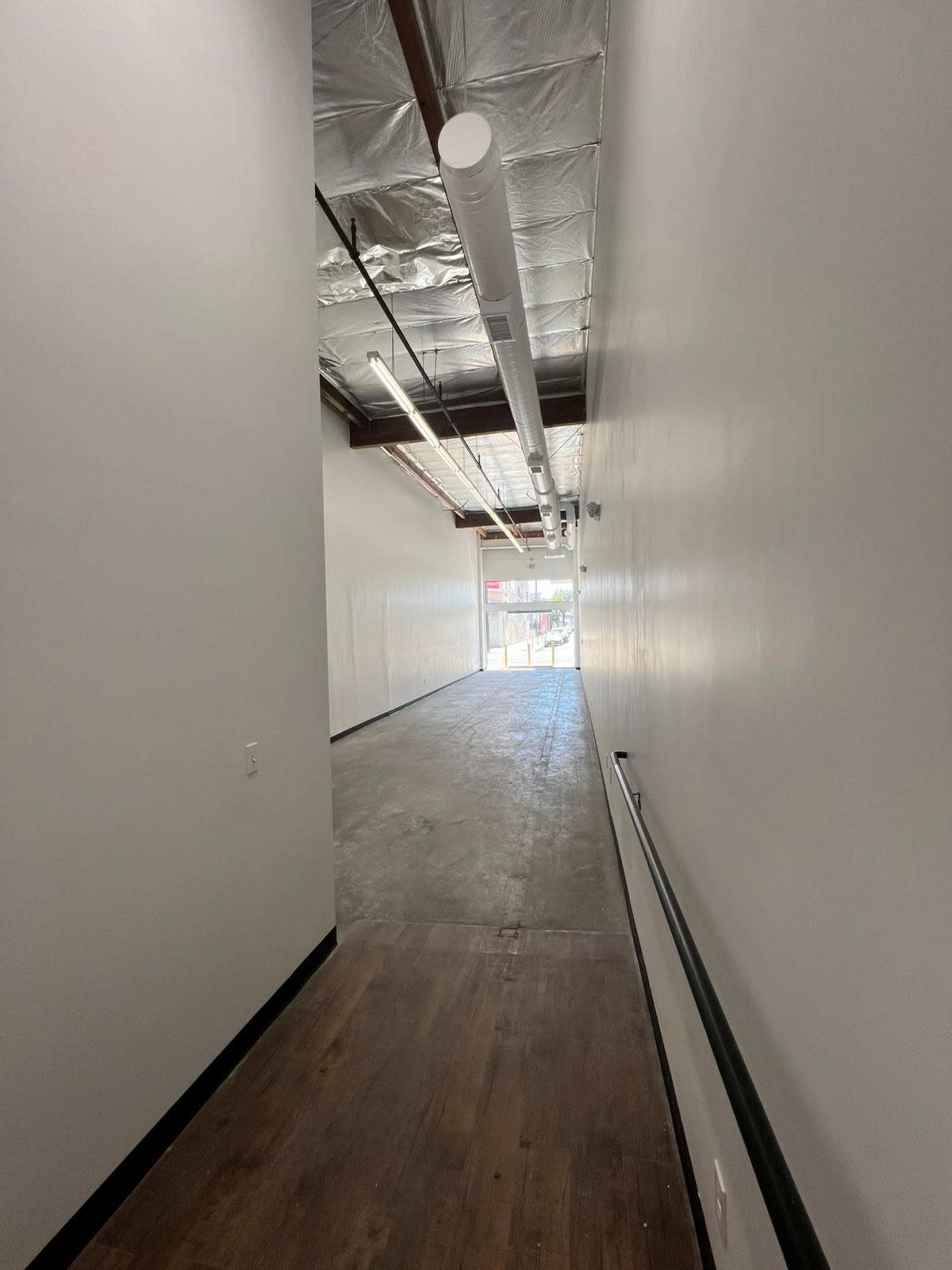 The image shows a long, narrow hallway with a smooth floor and white walls, leading to a bright entrance at the far end.