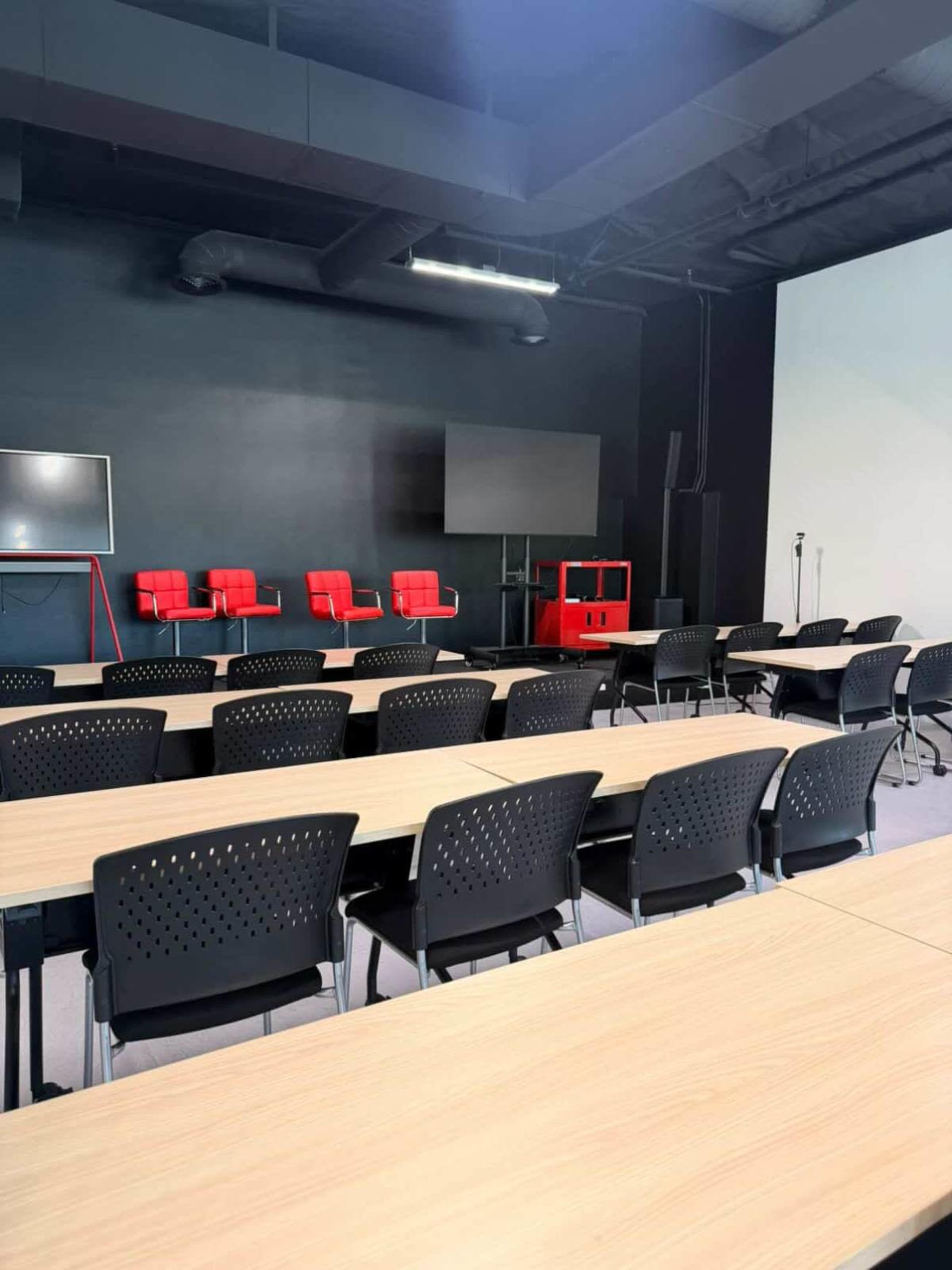 A conference room is set up with rows of black chairs and wooden tables facing a large screen and two red chairs at the front.