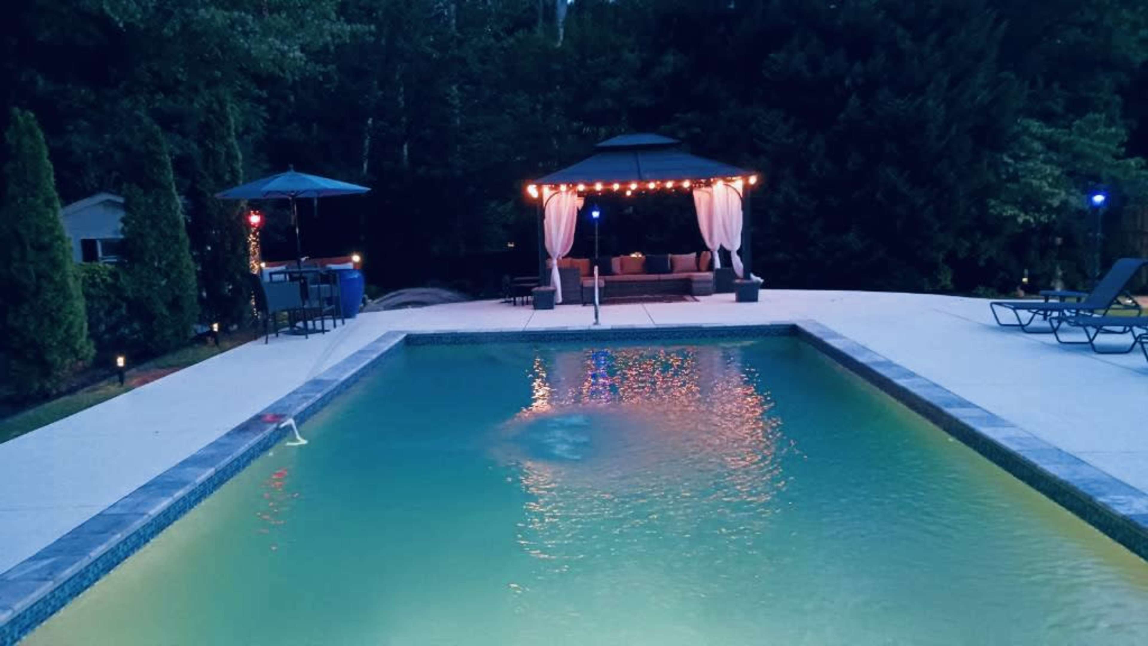 A pool surrounded by a patio area with a gazebo and lounge chairs, all illuminated by soft lighting in the evening.