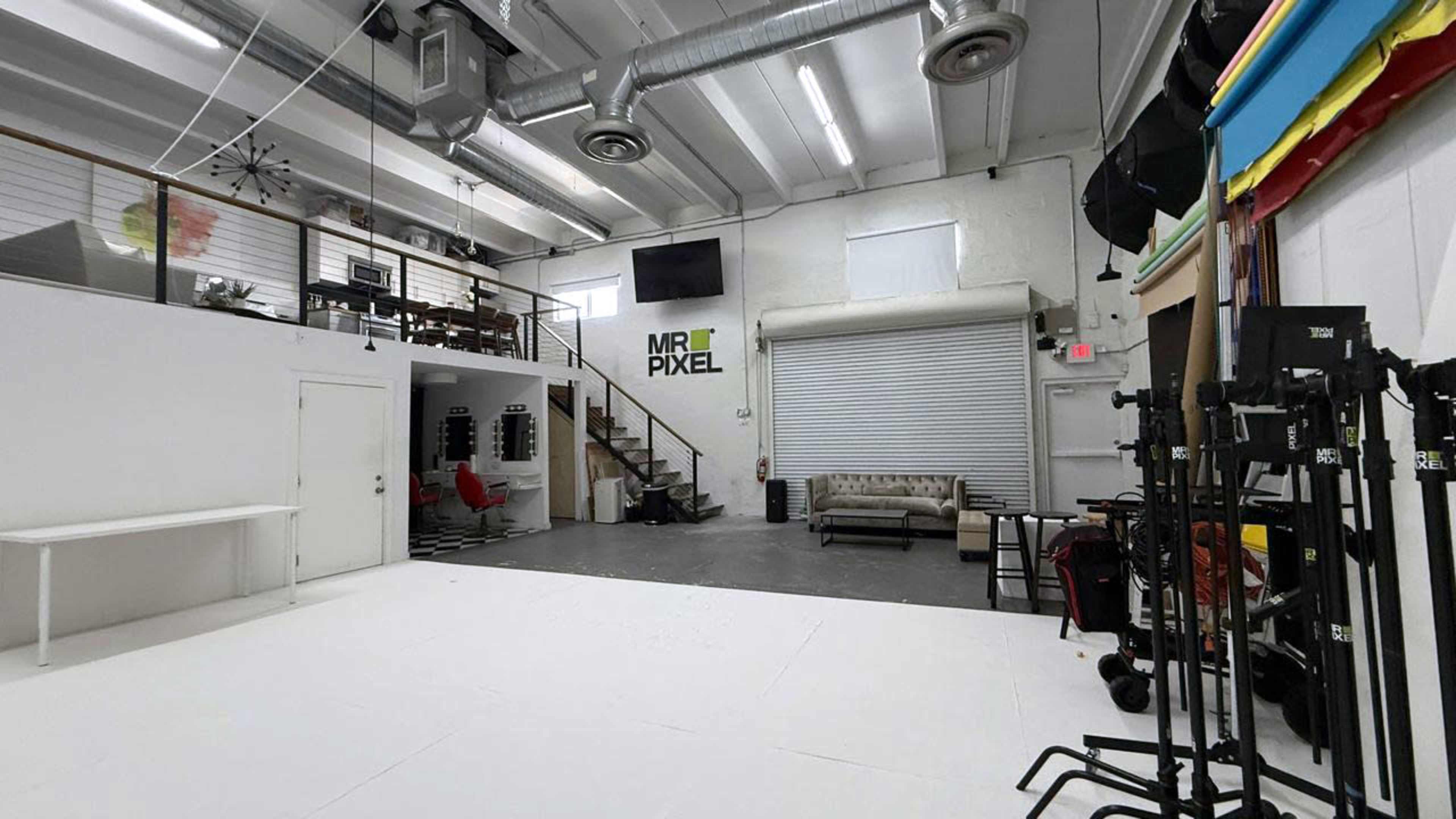 The image shows a spacious indoor studio with a white floor, a staircase leading to an upper level, and various equipment placed throughout the area.