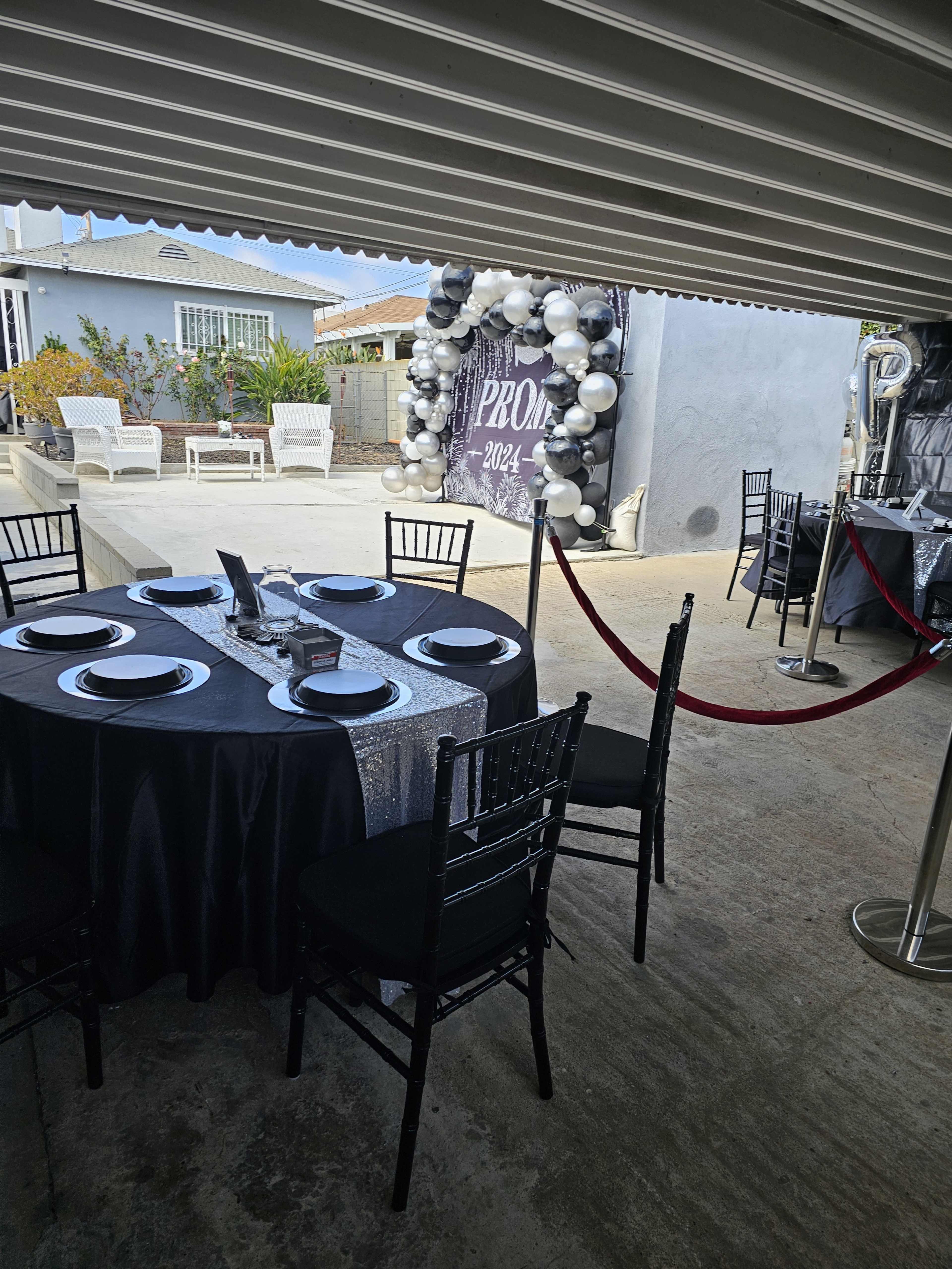 A round black table is set for an event, surrounded by black chairs, with a decorative archway of balloons in the background.