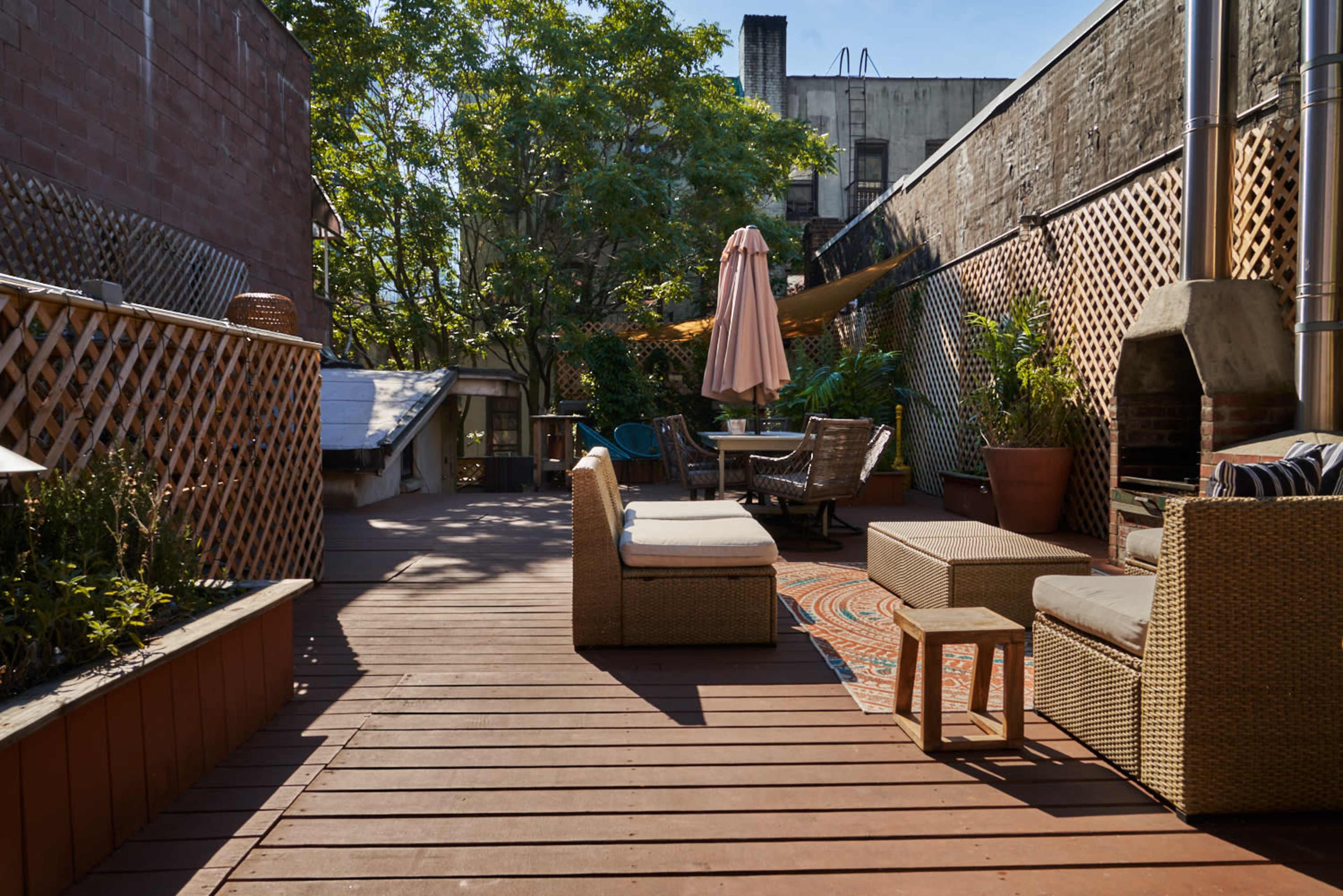 A wooden deck patio with outdoor furniture, plants, and an umbrella near a grill in an urban setting.