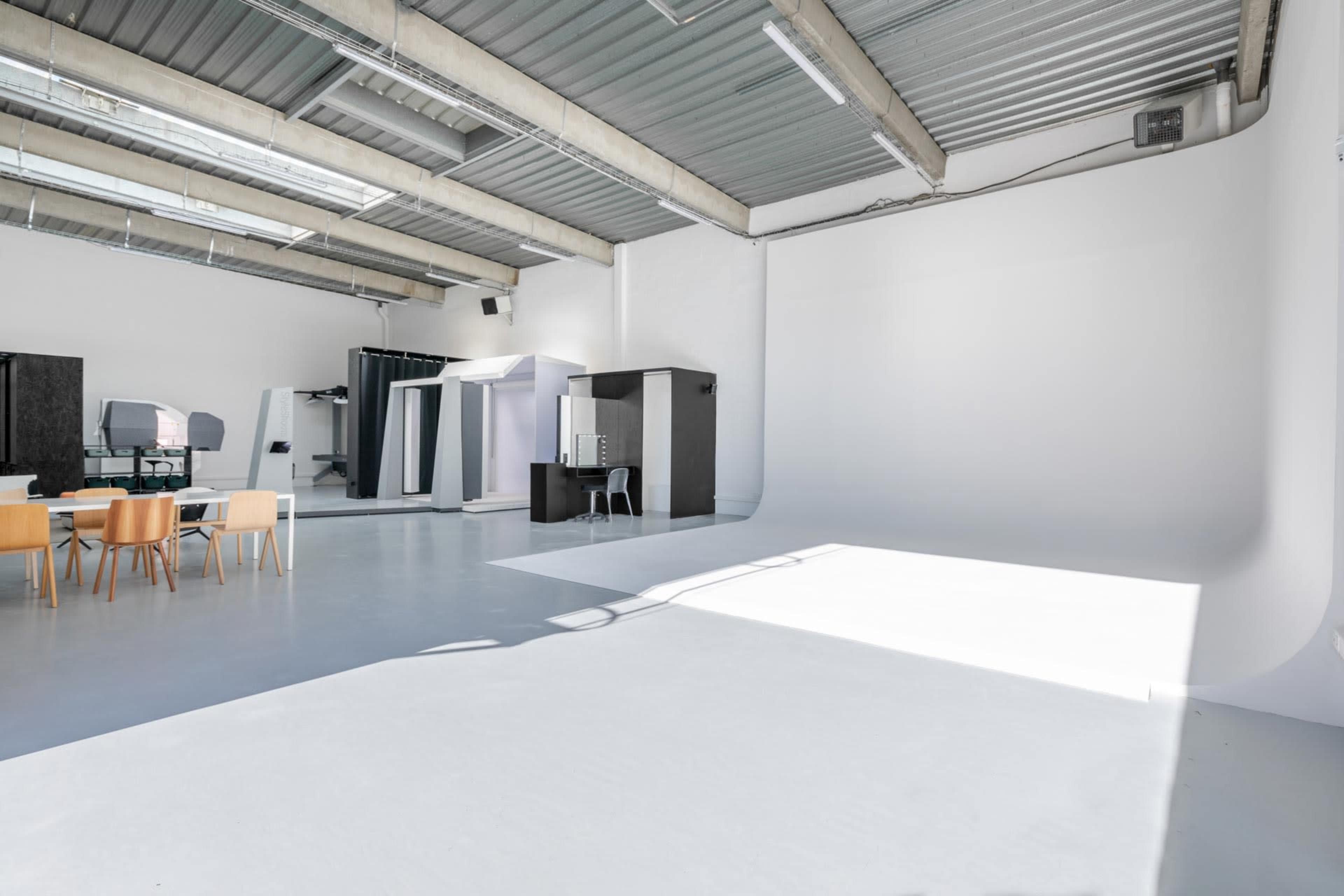 A bright, empty studio space featuring a white backdrop, a dining table with four chairs, and various furnishings against the walls.