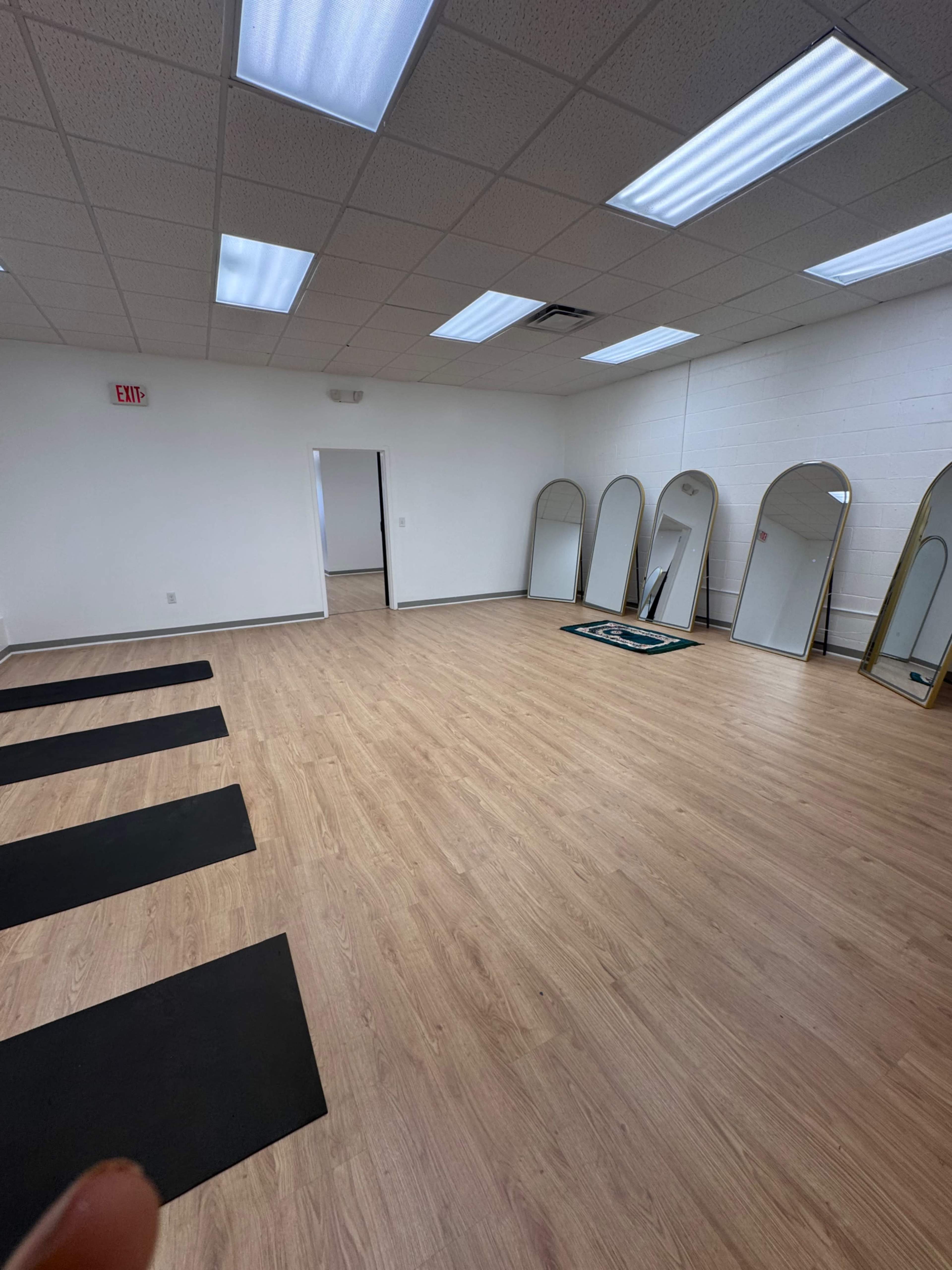 The image shows an empty room with light wood flooring, several yoga mats on the floor, and multiple tall mirrors mounted on the walls.