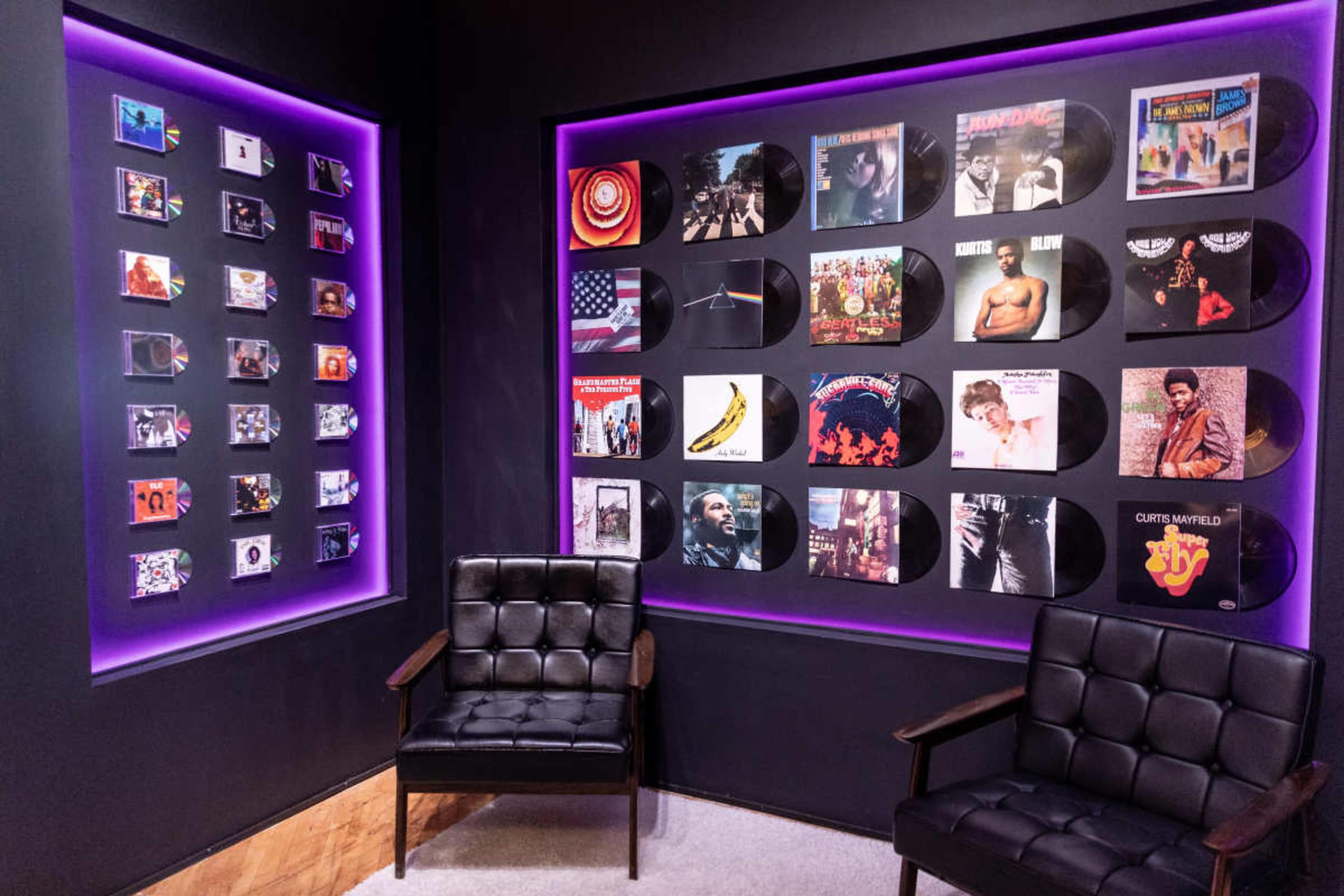 The image shows a stylish room with two black chairs facing a wall adorned with framed vinyl records and a smaller display of vintage media items, all highlighted by purple backlighting.