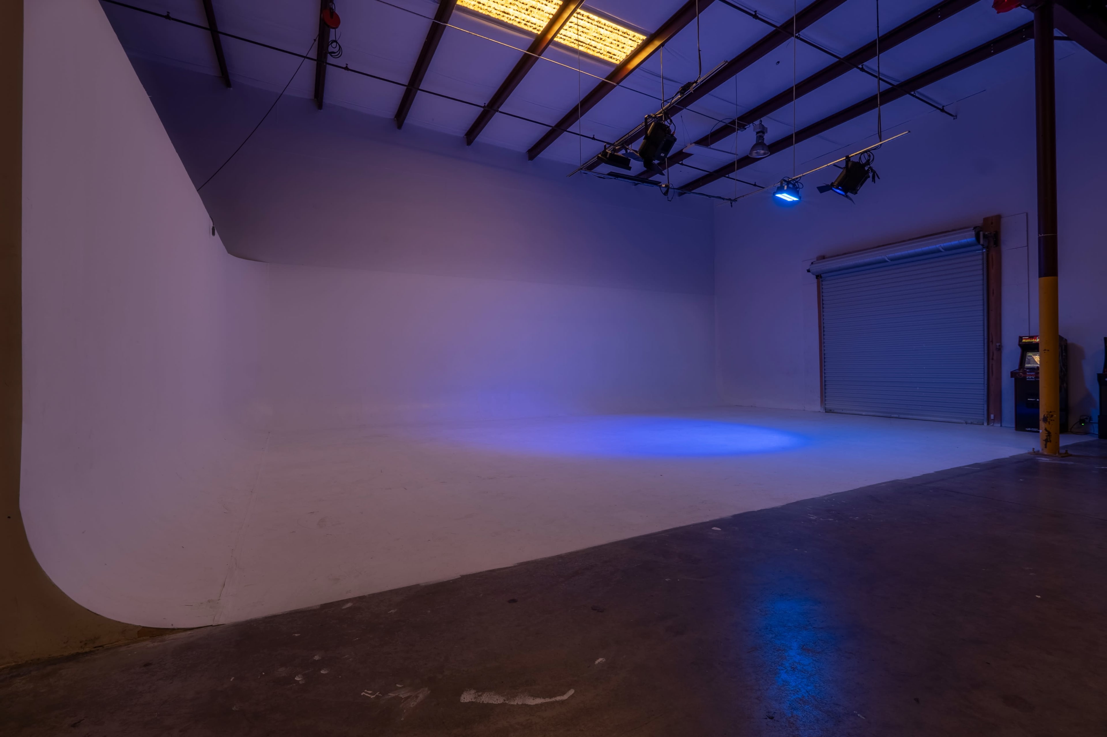 The image shows a spacious photography studio with a seamless cyclorama wall and a large roll-up door, illuminated by blue lighting.