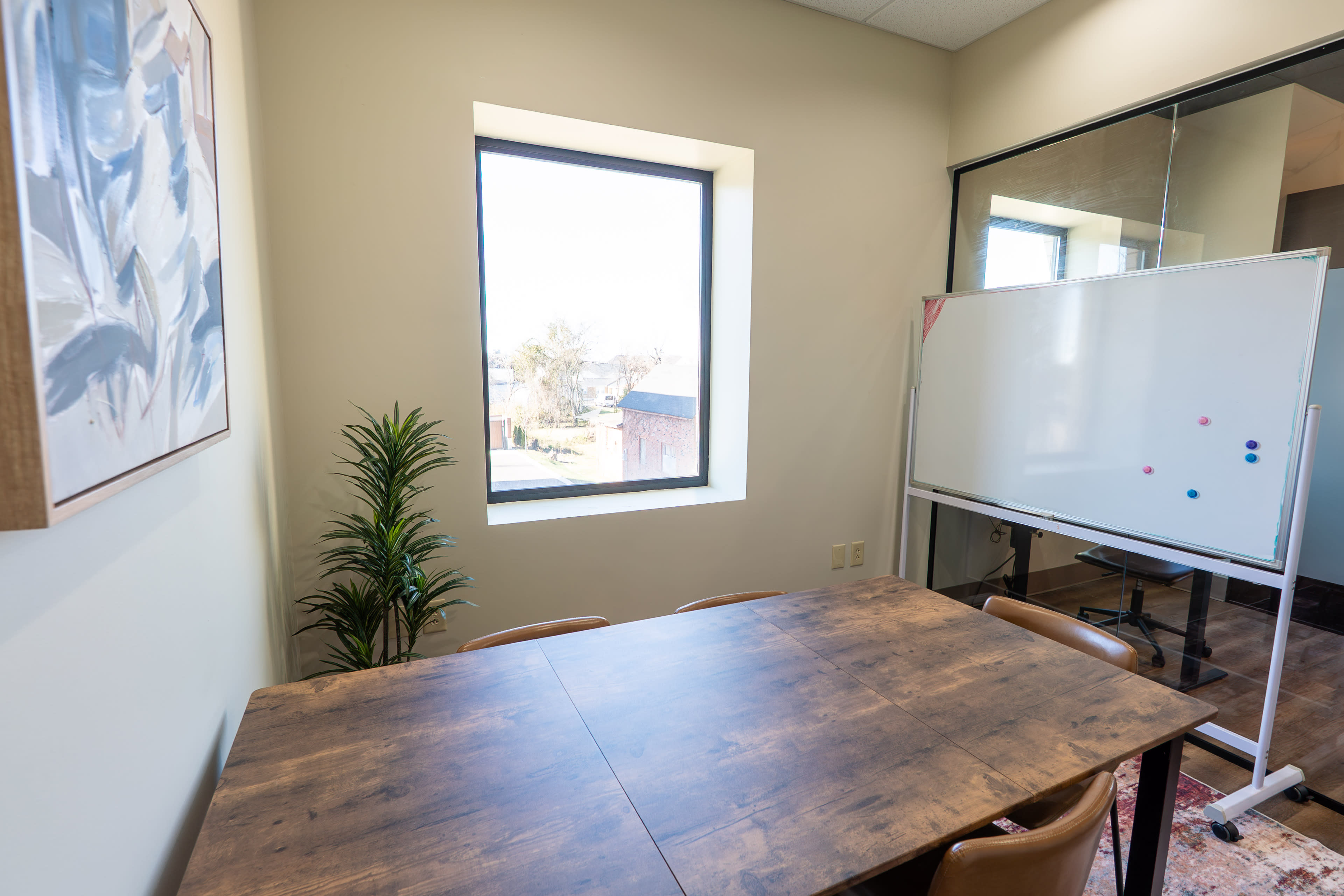 The image shows a small conference room with a wooden table, four chairs, a large window, a potted plant, and a whiteboard.