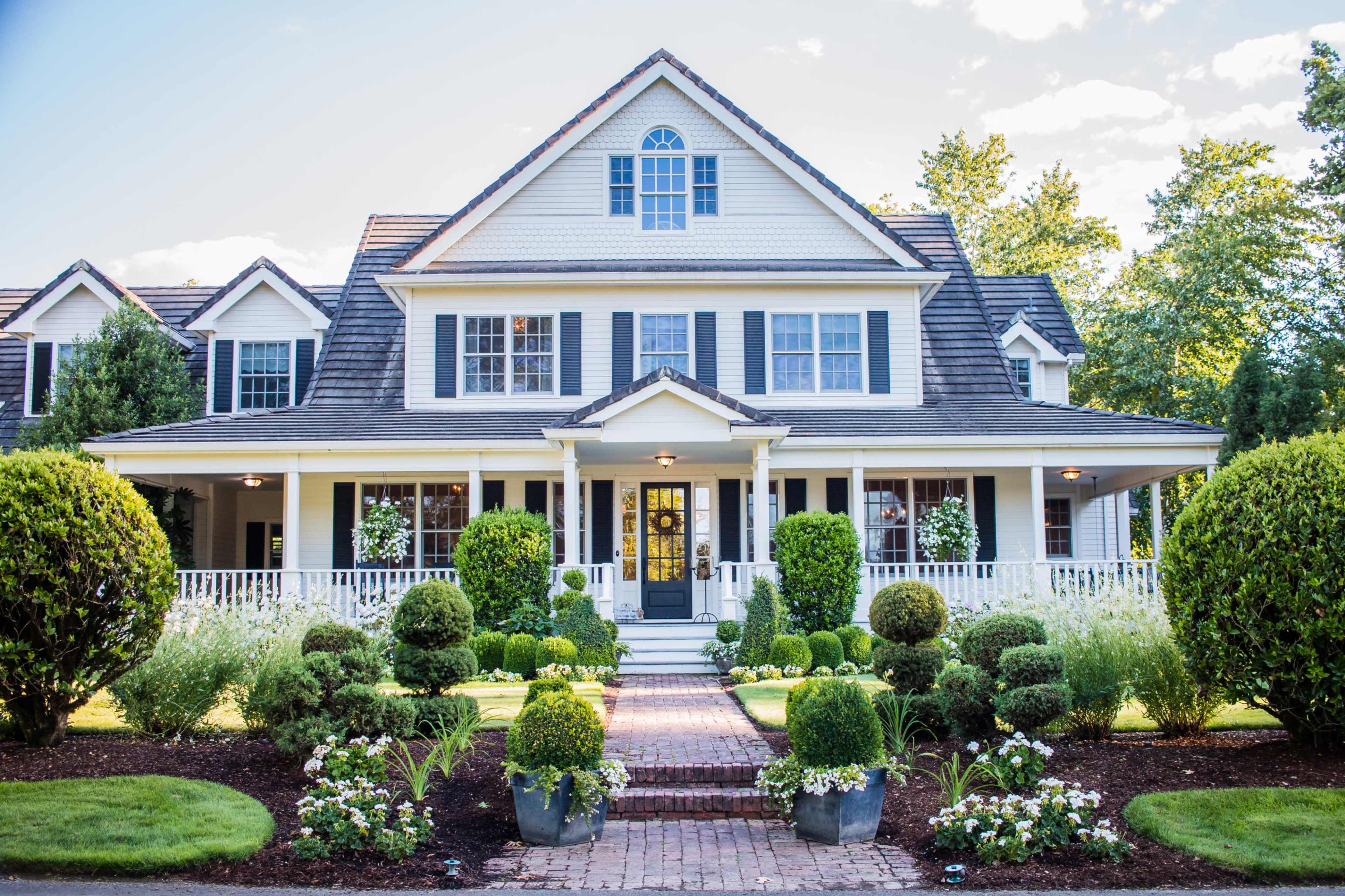 A large, two-story house with a steeply pitched roof and a front porch is surrounded by well-maintained shrubs and flowerbeds.