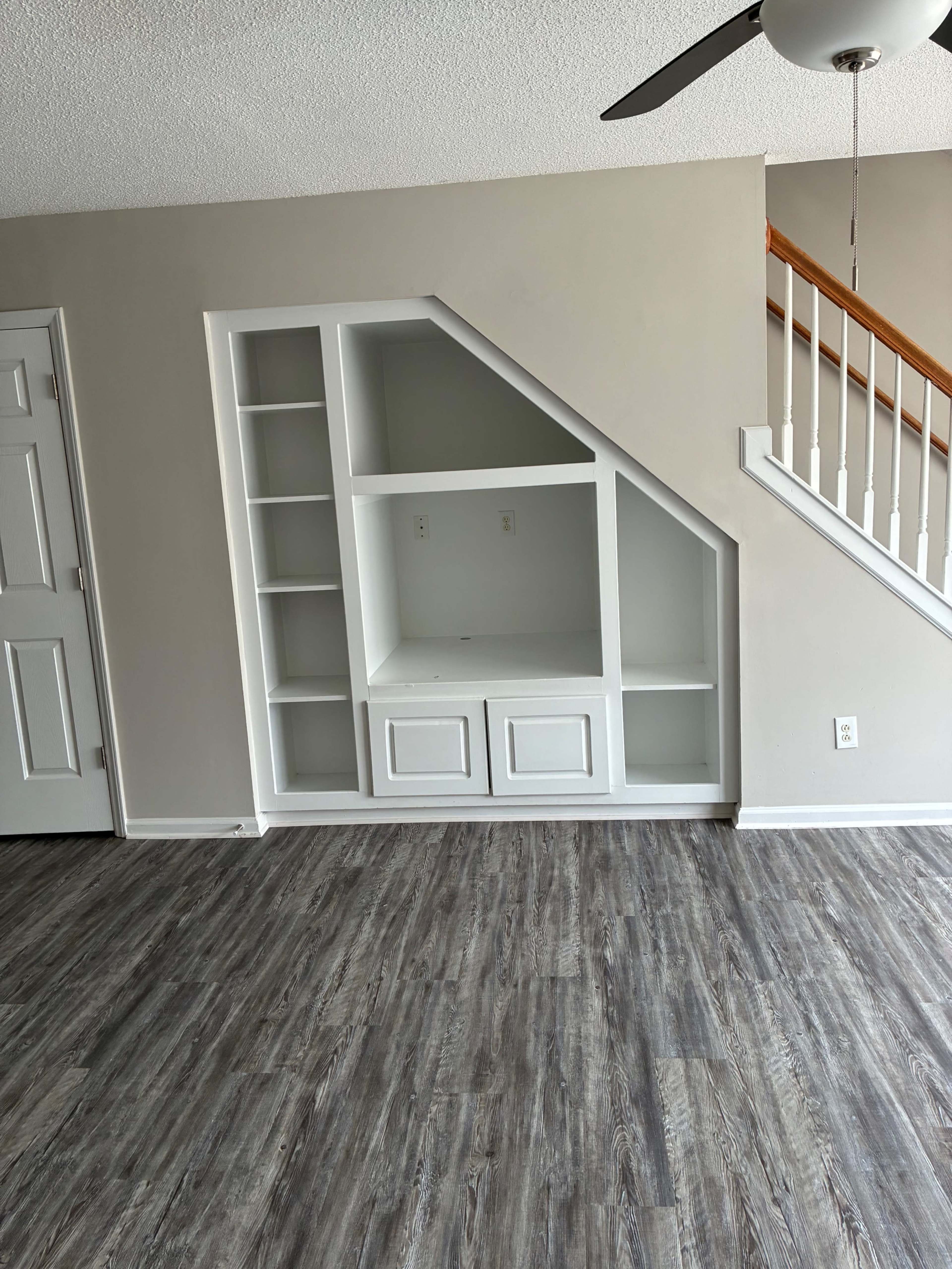 The image shows a corner built-in bookshelf unit with open shelves and a closed cabinet in a room with a light-colored wall and wooden staircase.