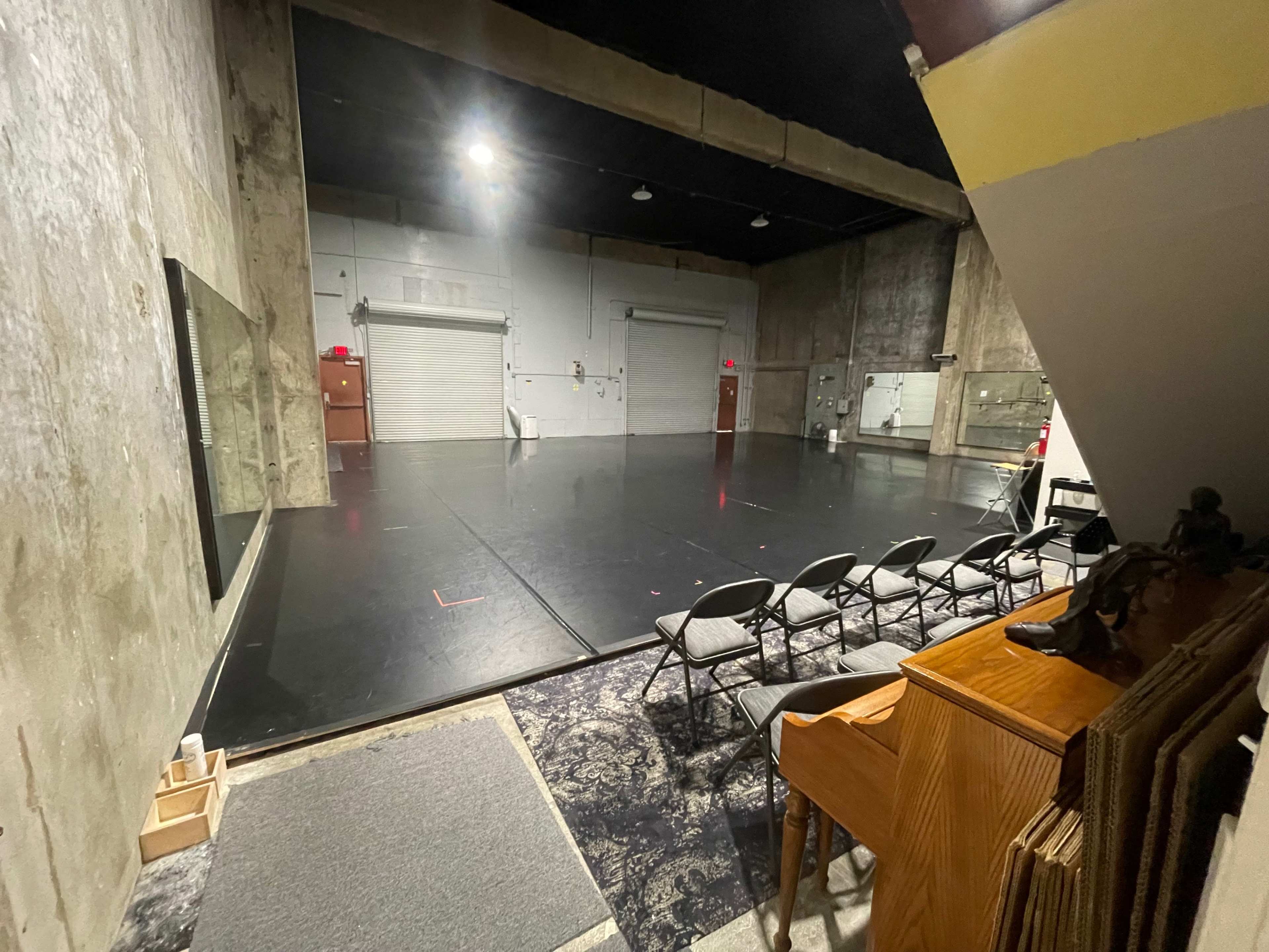 An empty rehearsal studio features a mirrored wall, a black floor, and folding chairs arranged in front of a small stage area.