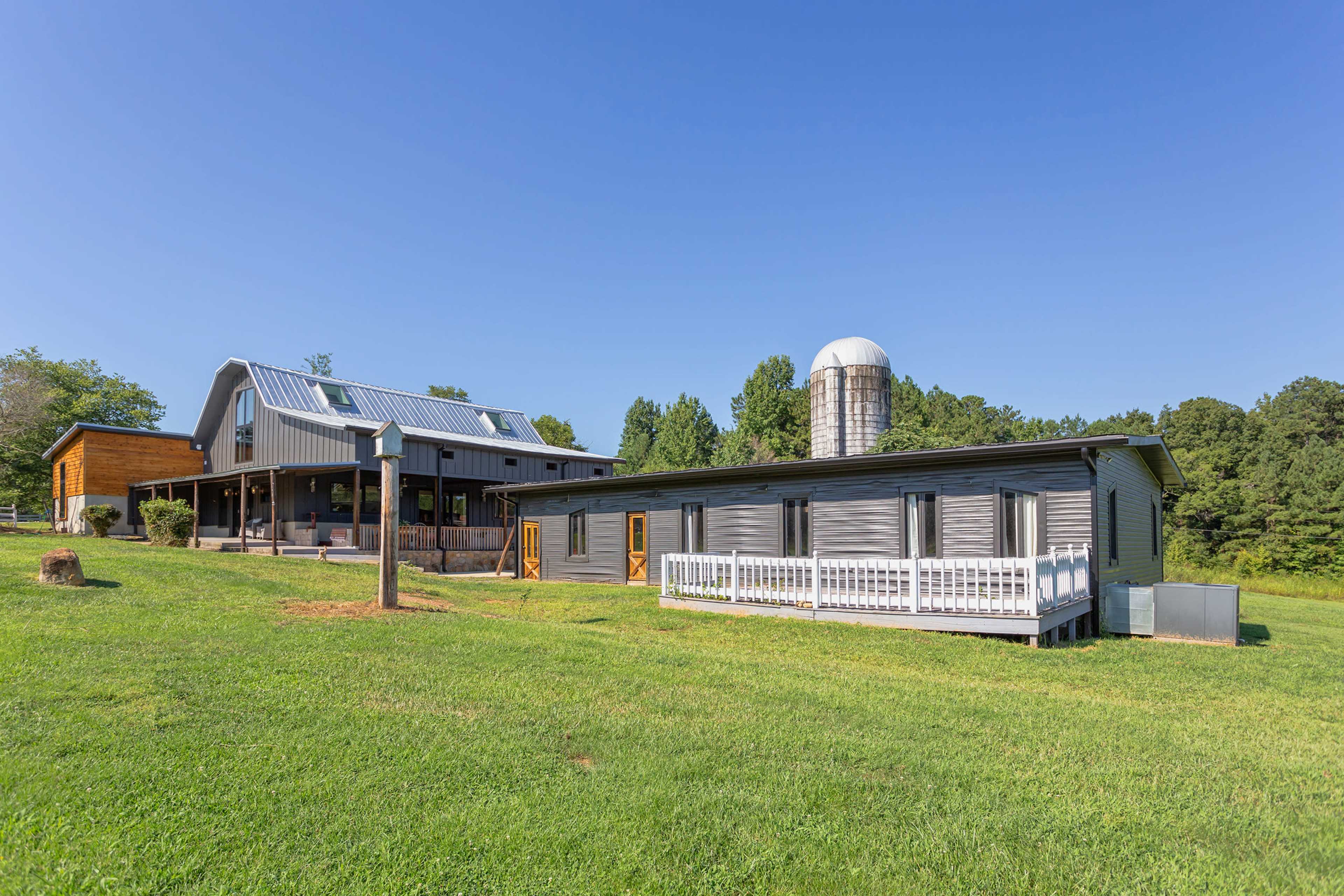 A modern farmhouse with a metal roof and a silos sits on a grassy slope surrounded by trees.