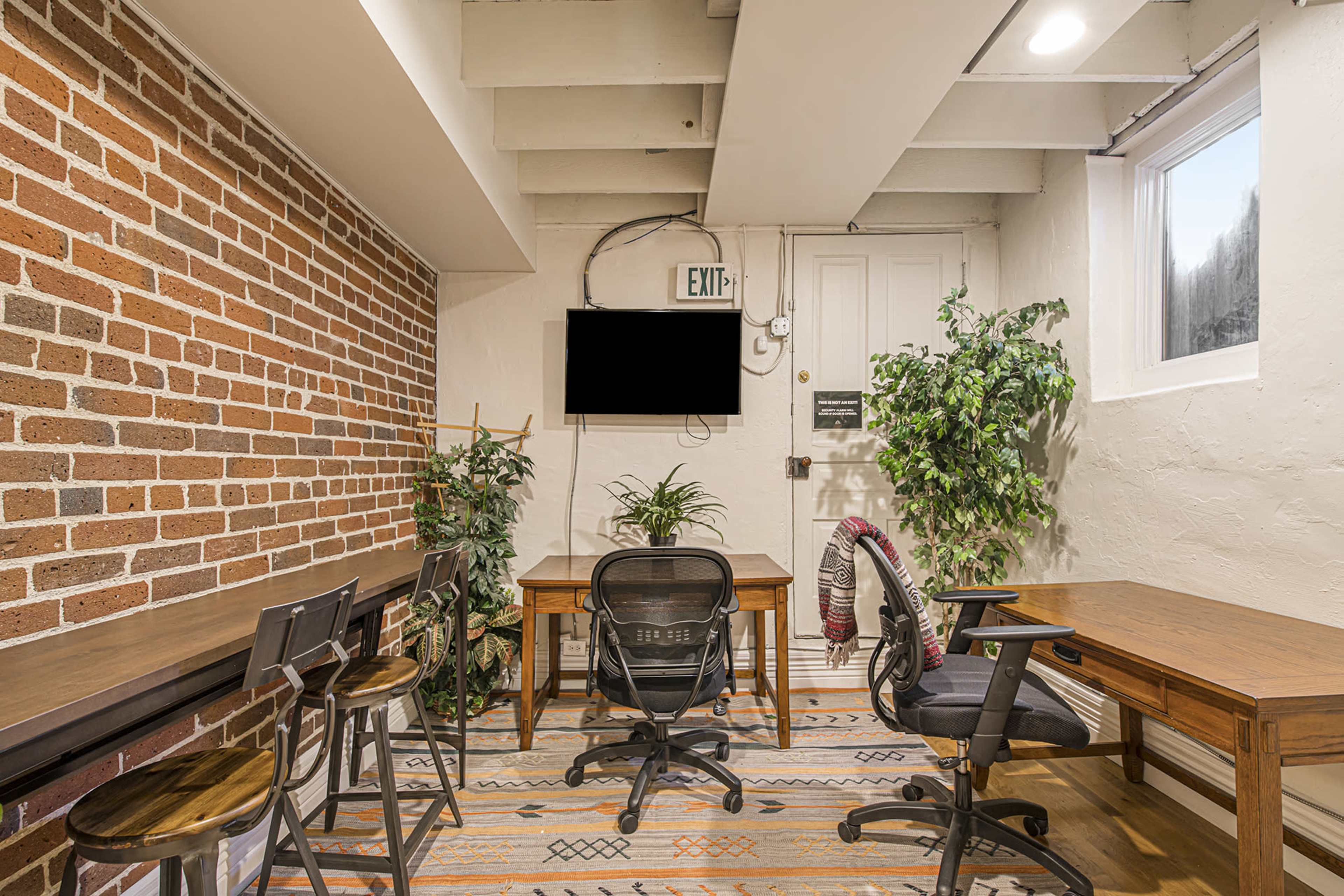 A small office space with brick walls, wooden tables, chairs, indoor plants, and a wall-mounted television.
