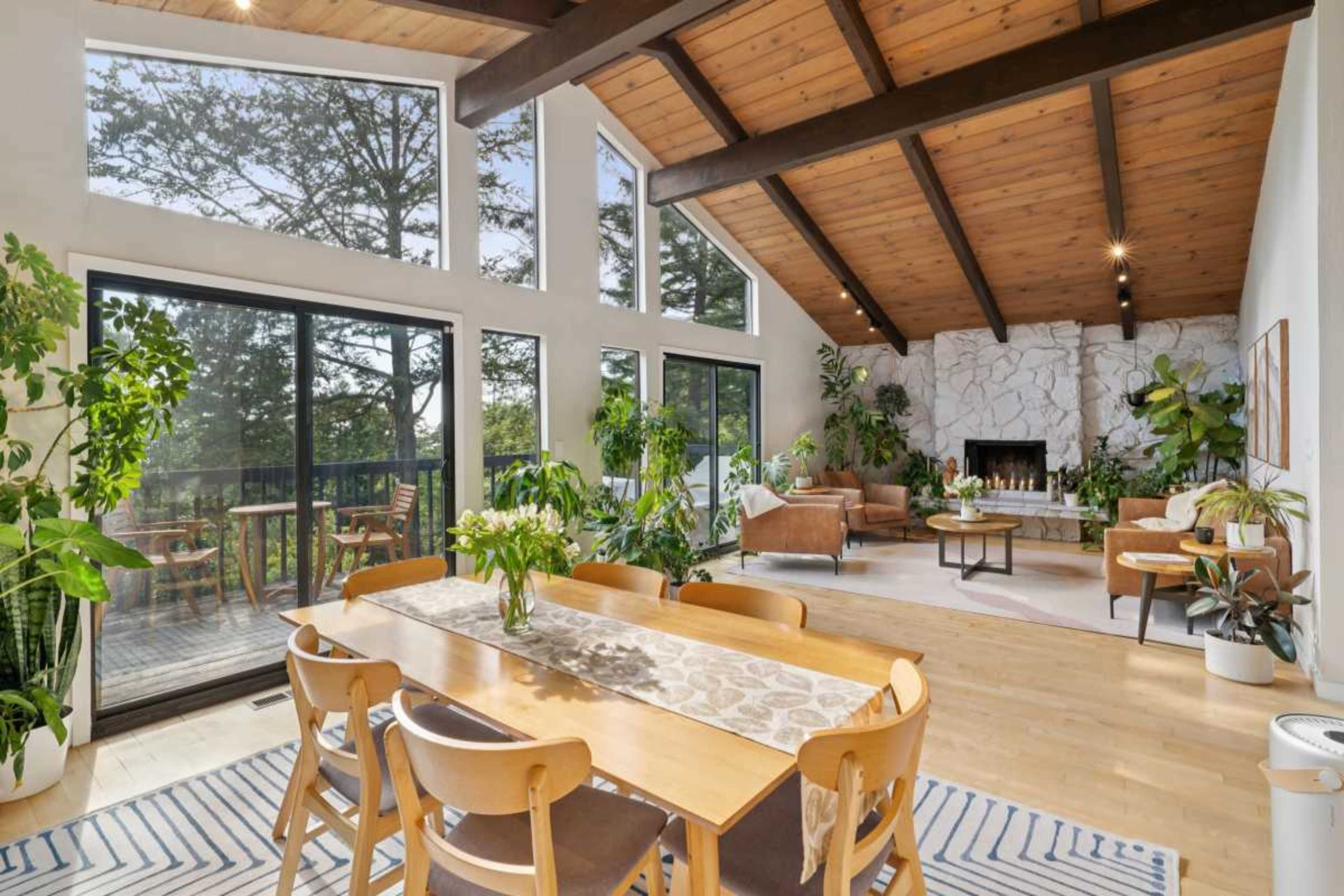 The image shows a spacious living area with large windows, a wooden dining table, and various indoor plants, featuring a stone fireplace and a deck visible through the sliding glass doors.