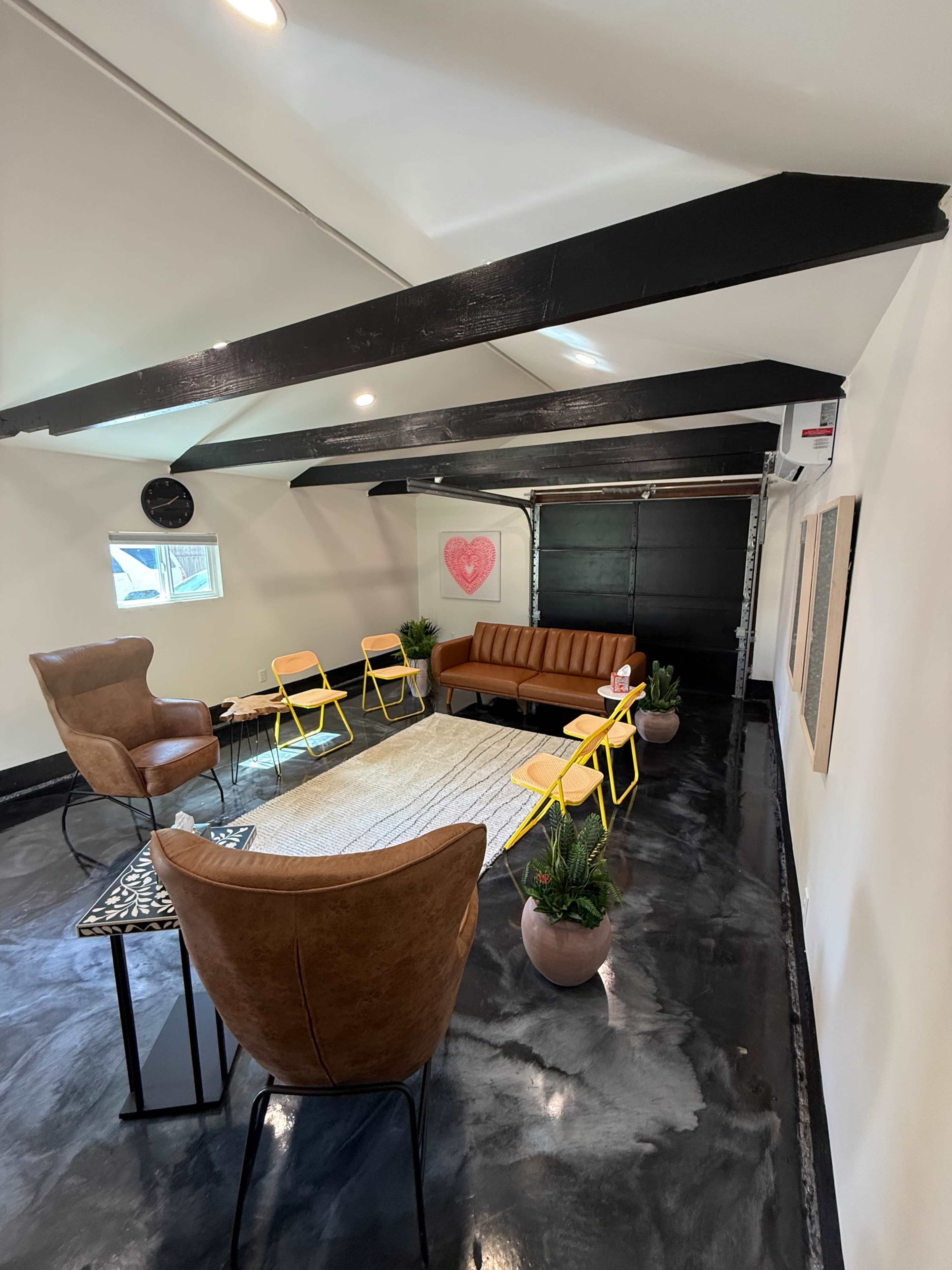 The image shows a modern interior space with a leather sofa, two armchairs, yellow chairs, and a rug, arranged around a coffee table beneath wooden beams.