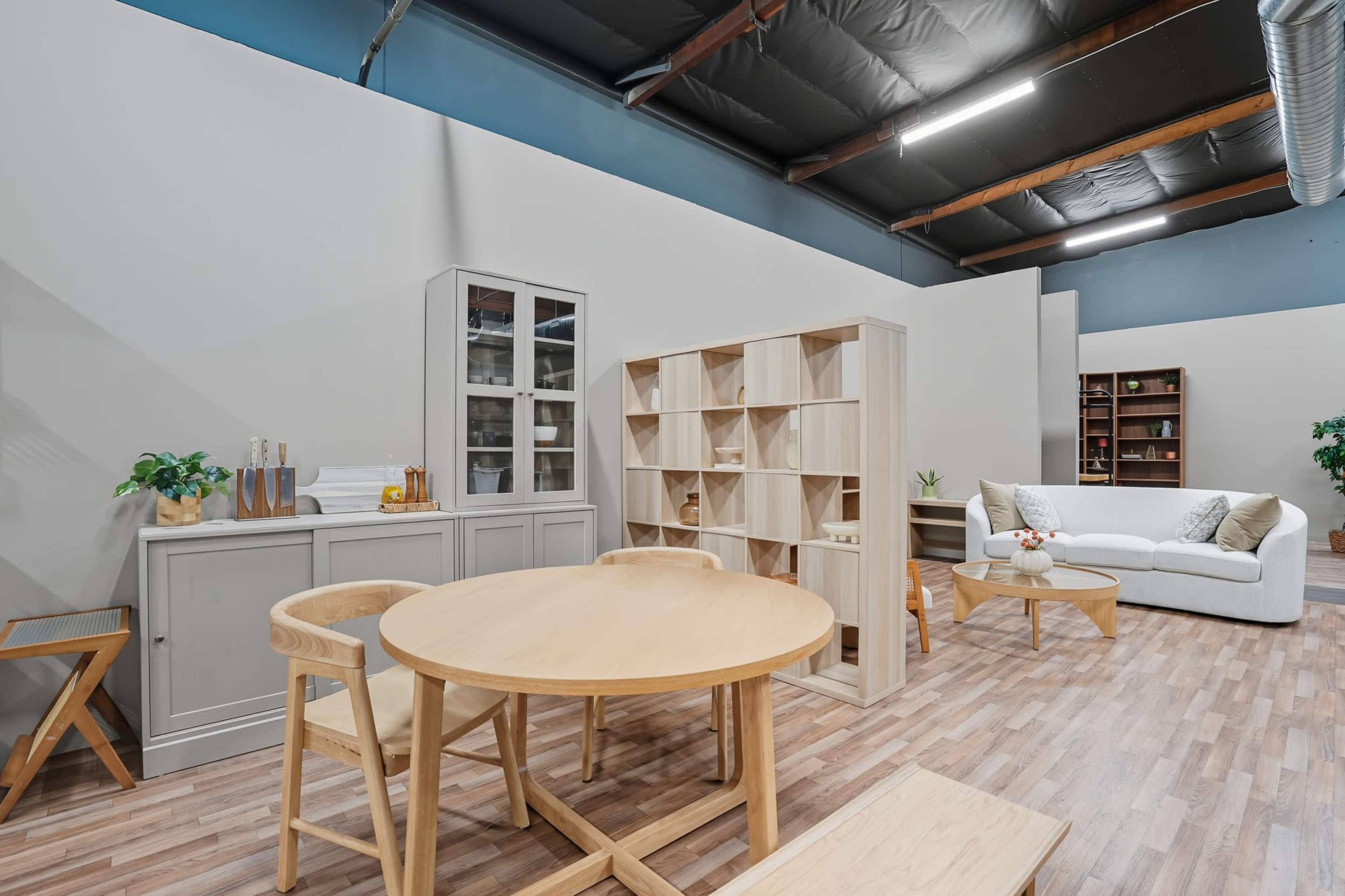 The image shows a spacious interior with wooden furniture, including a round dining table with chairs, a modular shelving unit, and a light-colored sofa in a well-lit room.
