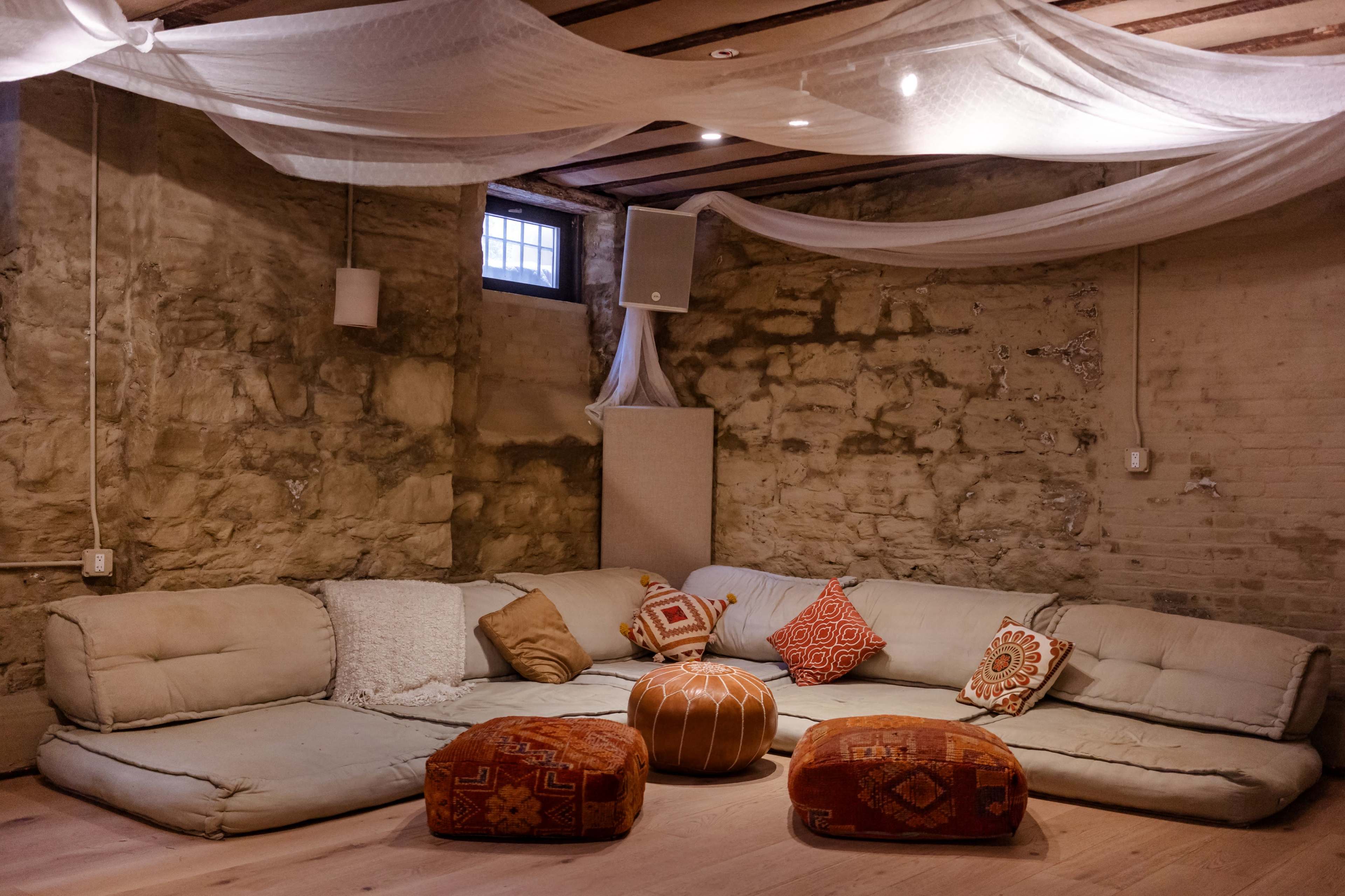 The image shows a cozy lounge area with a large, U-shaped seating arrangement, decorated with colorful cushions and a round wooden table in the center.