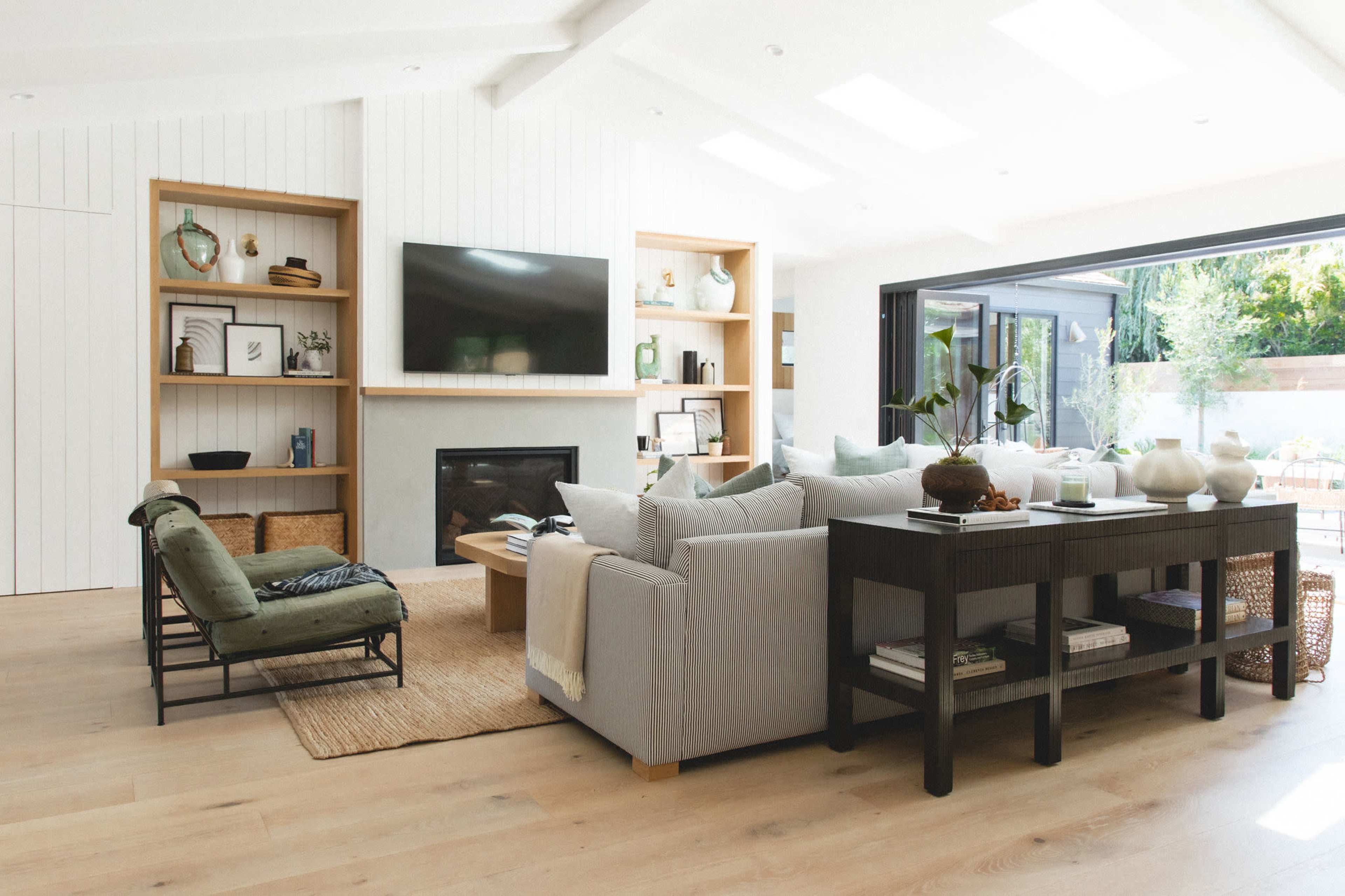 The living room features a large flat-screen TV mounted above a fireplace, surrounded by built-in shelves, a sectional sofa, and a console table, with large windows providing natural light.