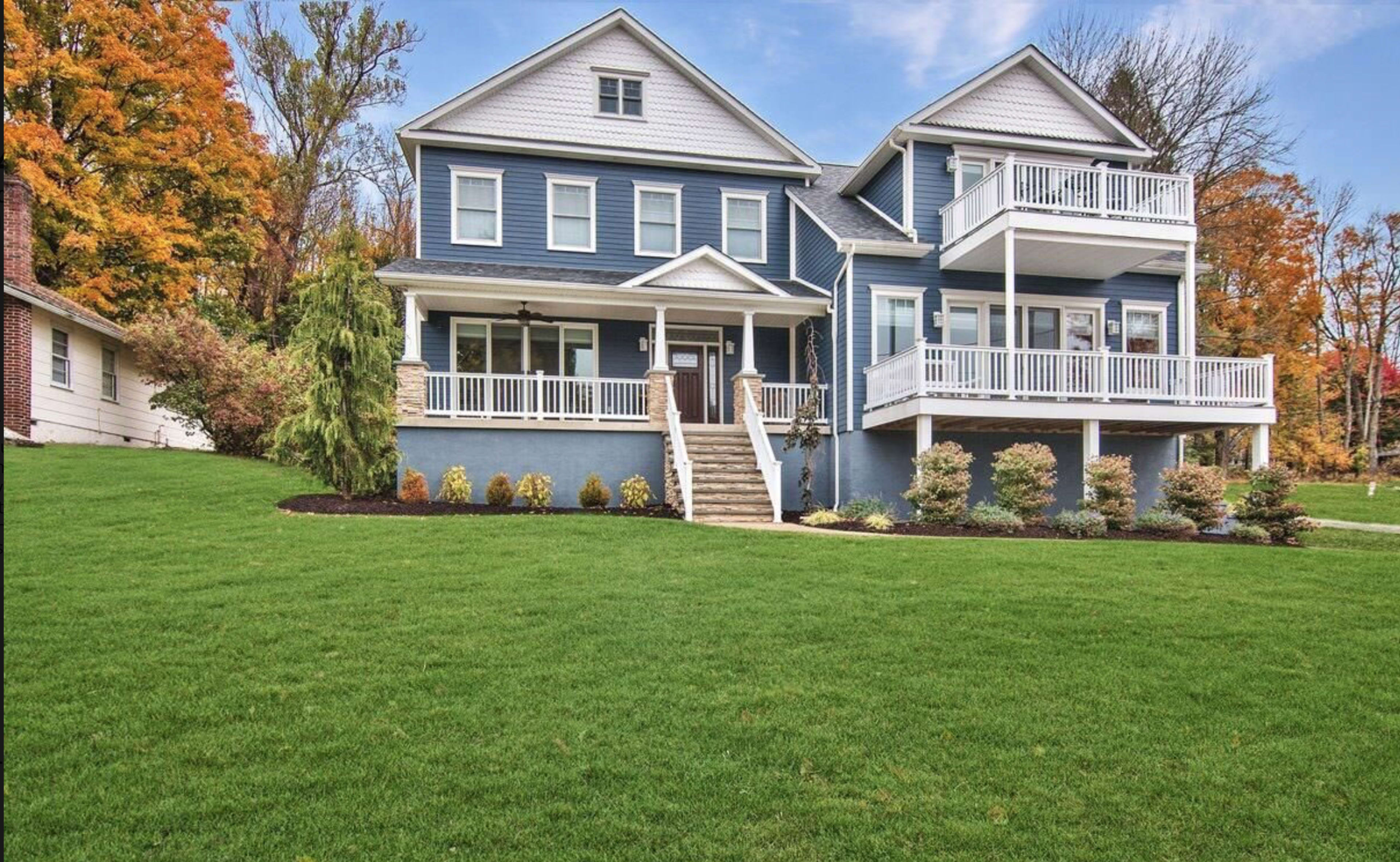 A two-story blue house with white trim and multiple balconies, surrounded by a well-maintained lawn and landscaped shrubs.