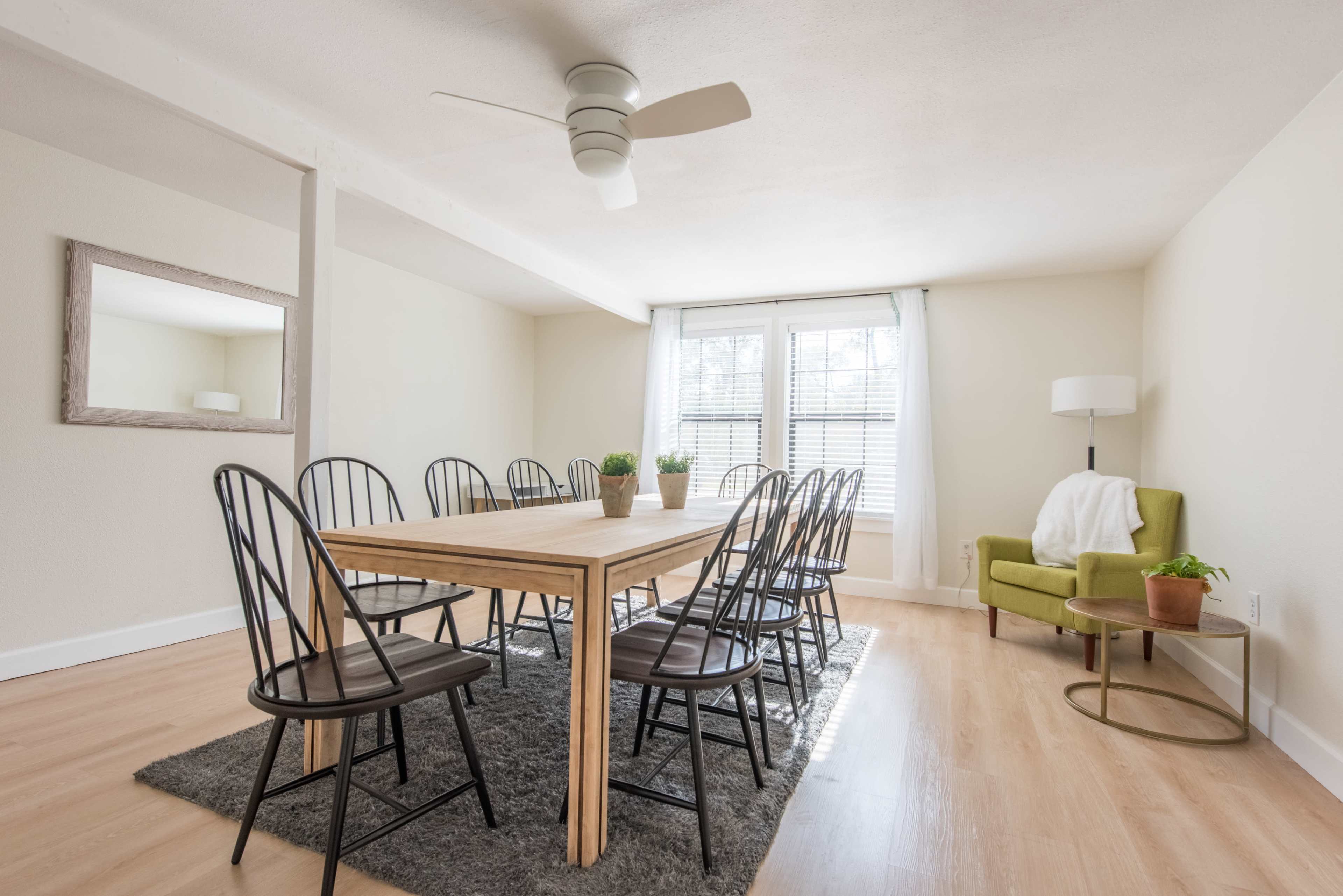 A dining room features a large wooden table surrounded by black chairs, with a green armchair in one corner and a ceiling fan overhead.