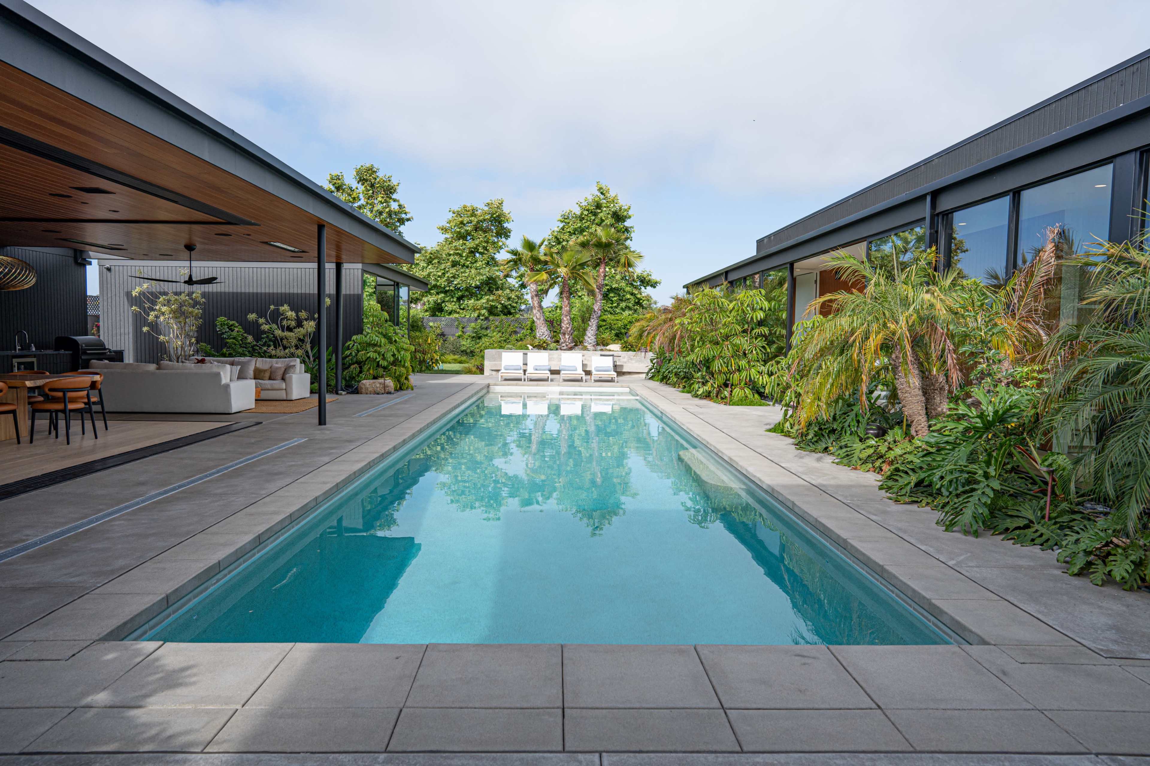 A modern outdoor area features a rectangular swimming pool surrounded by tropical plants and lounge chairs.
