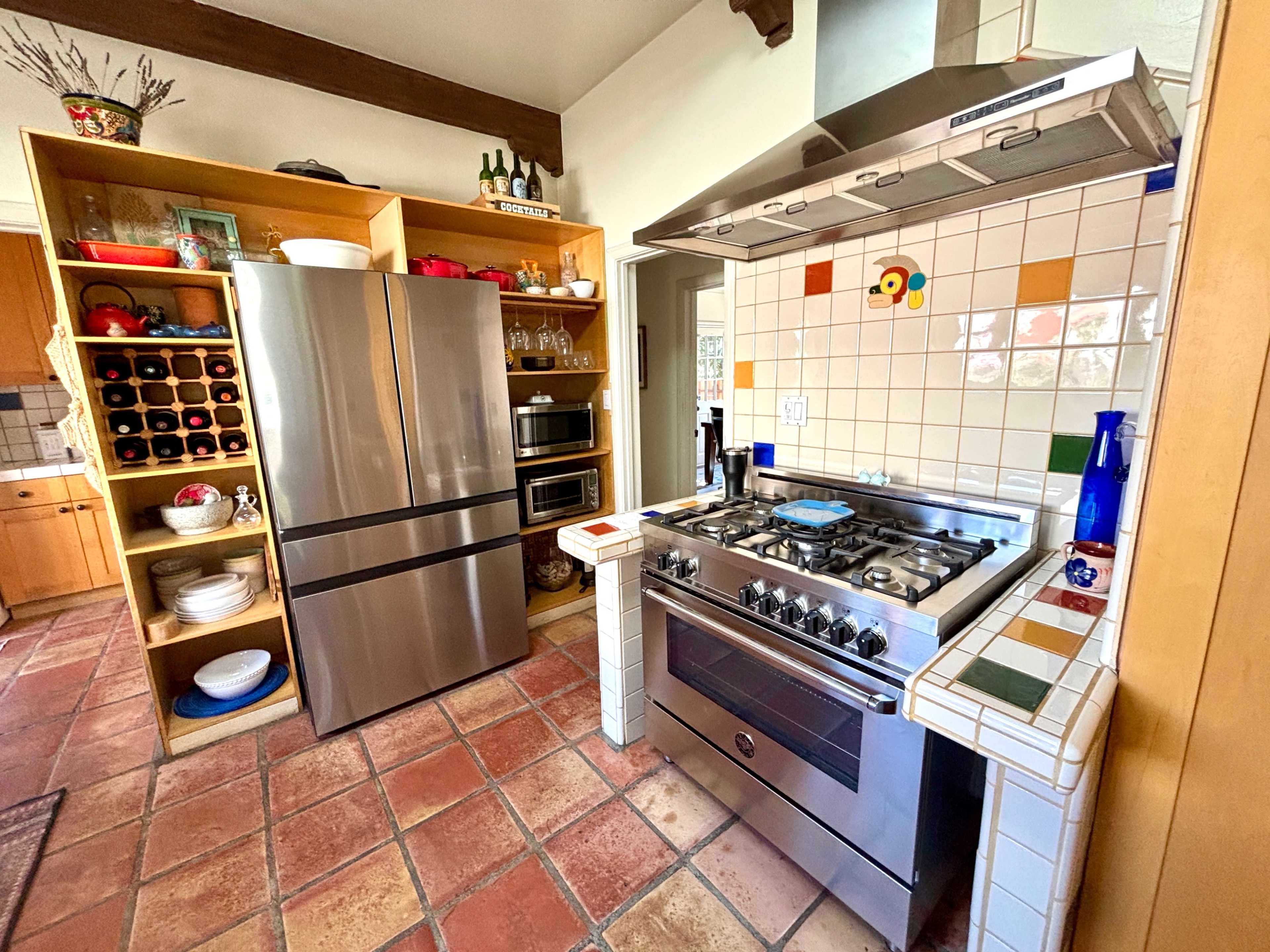 The image shows a kitchen featuring stainless steel appliances, including a refrigerator and a gas stove, with terracotta tiled flooring and wooden shelving displaying dishware and wine bottles.
