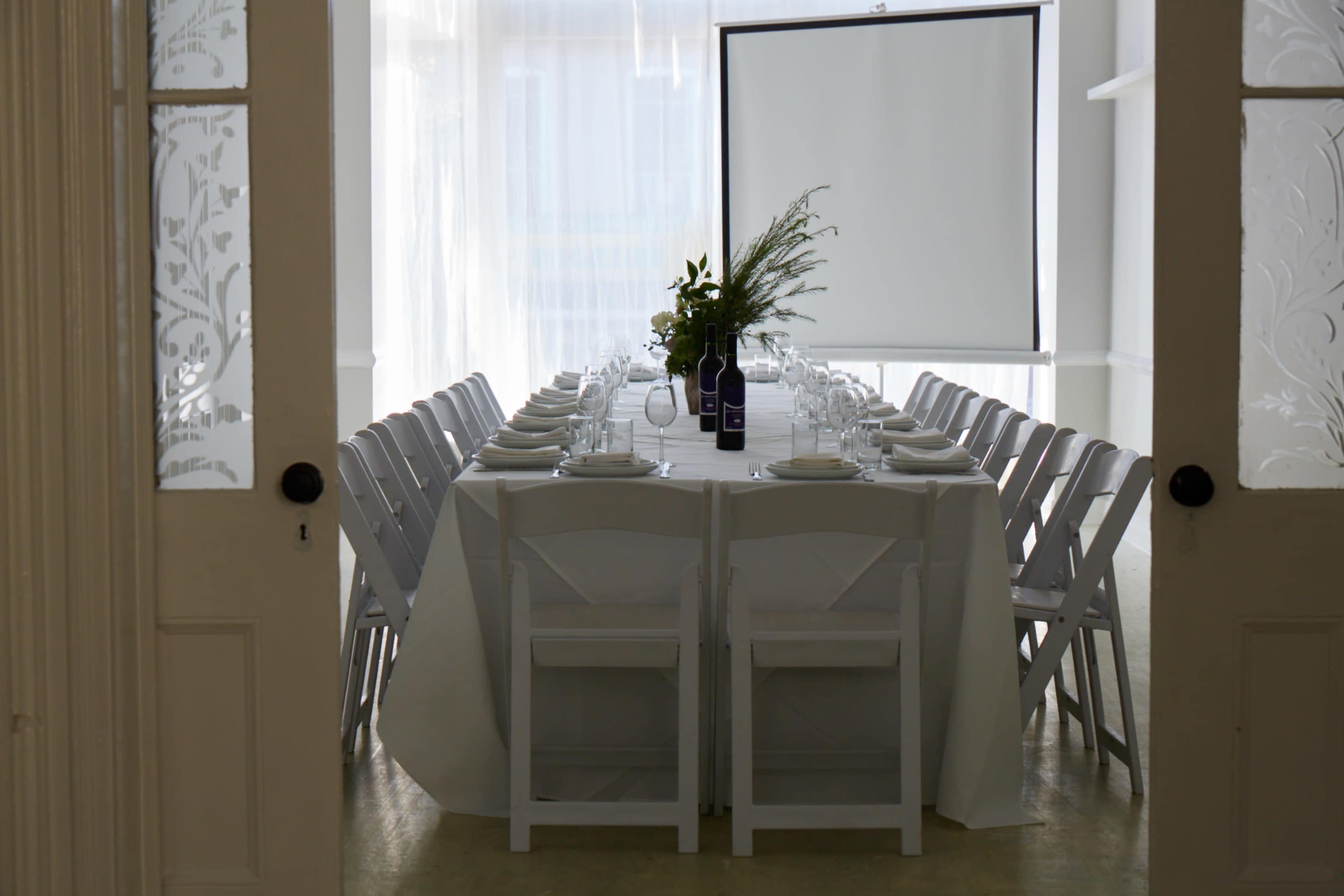 A long dining table is set with white plates and glasses, surrounded by white chairs in a bright, minimalistic room.