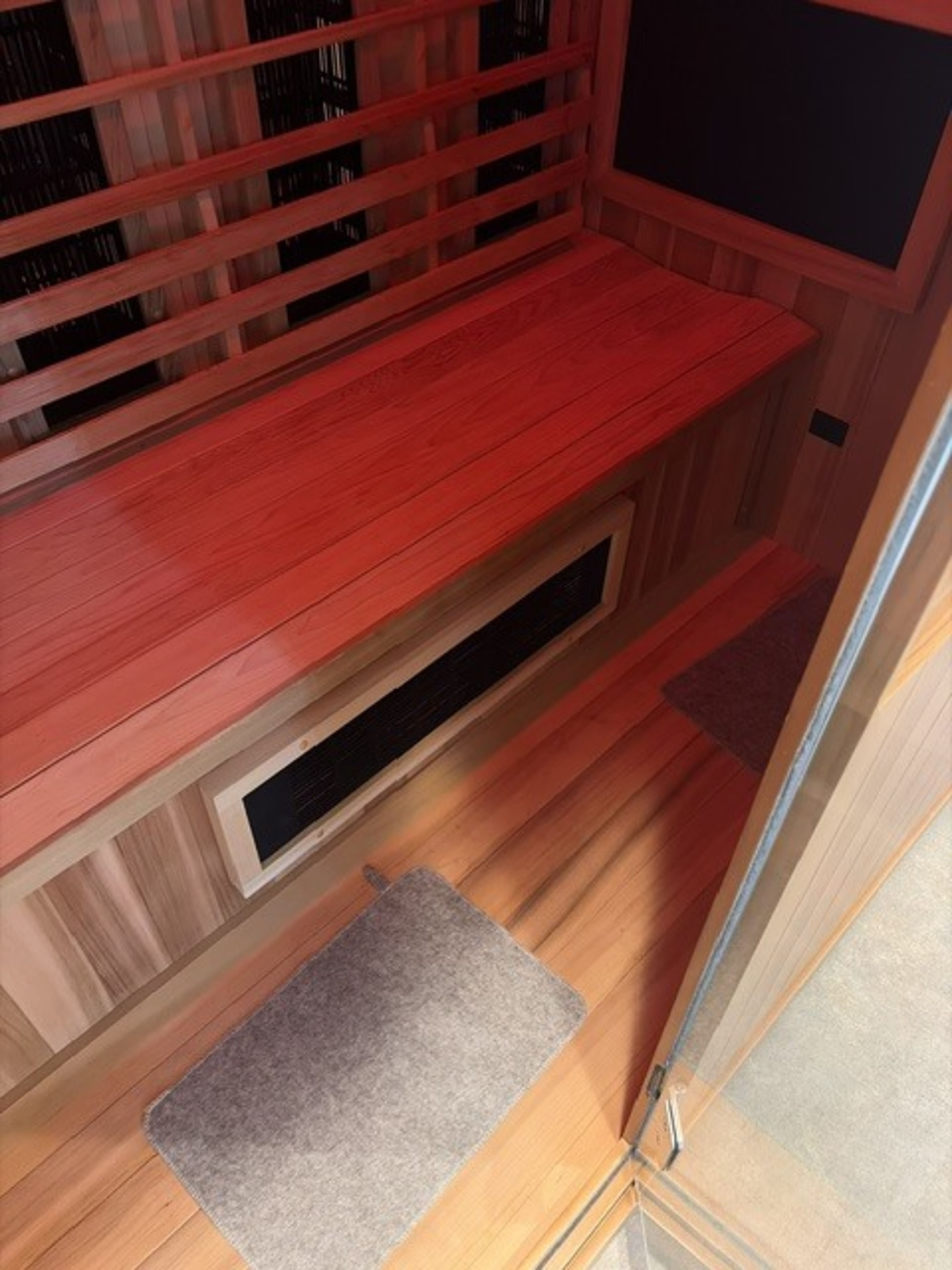 The image shows the interior of a wooden sauna featuring benches, a heater vent, and two small gray mats on the floor.