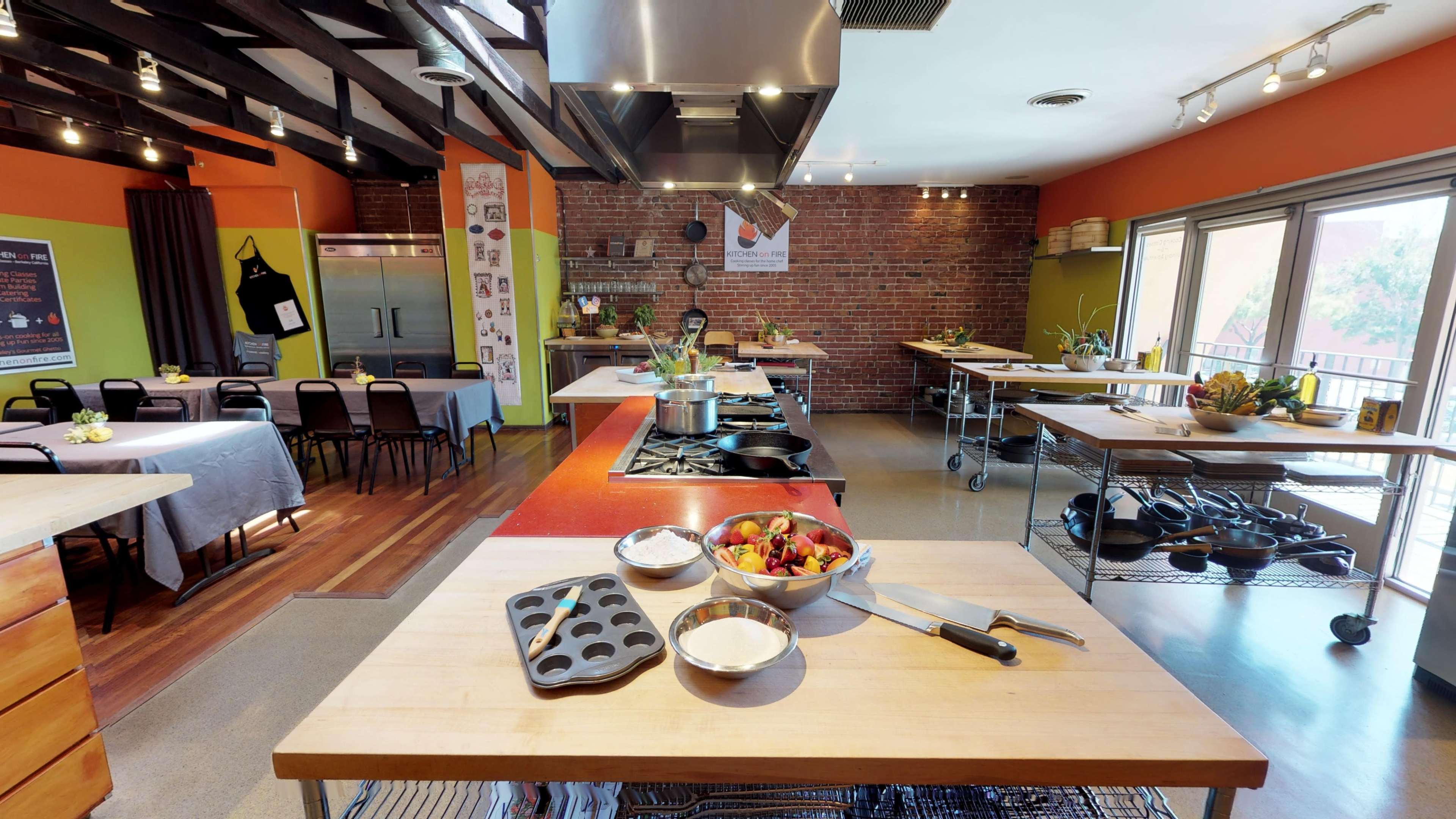 The image shows a spacious kitchen with multiple workstations, equipped with cooking appliances and ingredients, and features exposed brick walls and large windows.