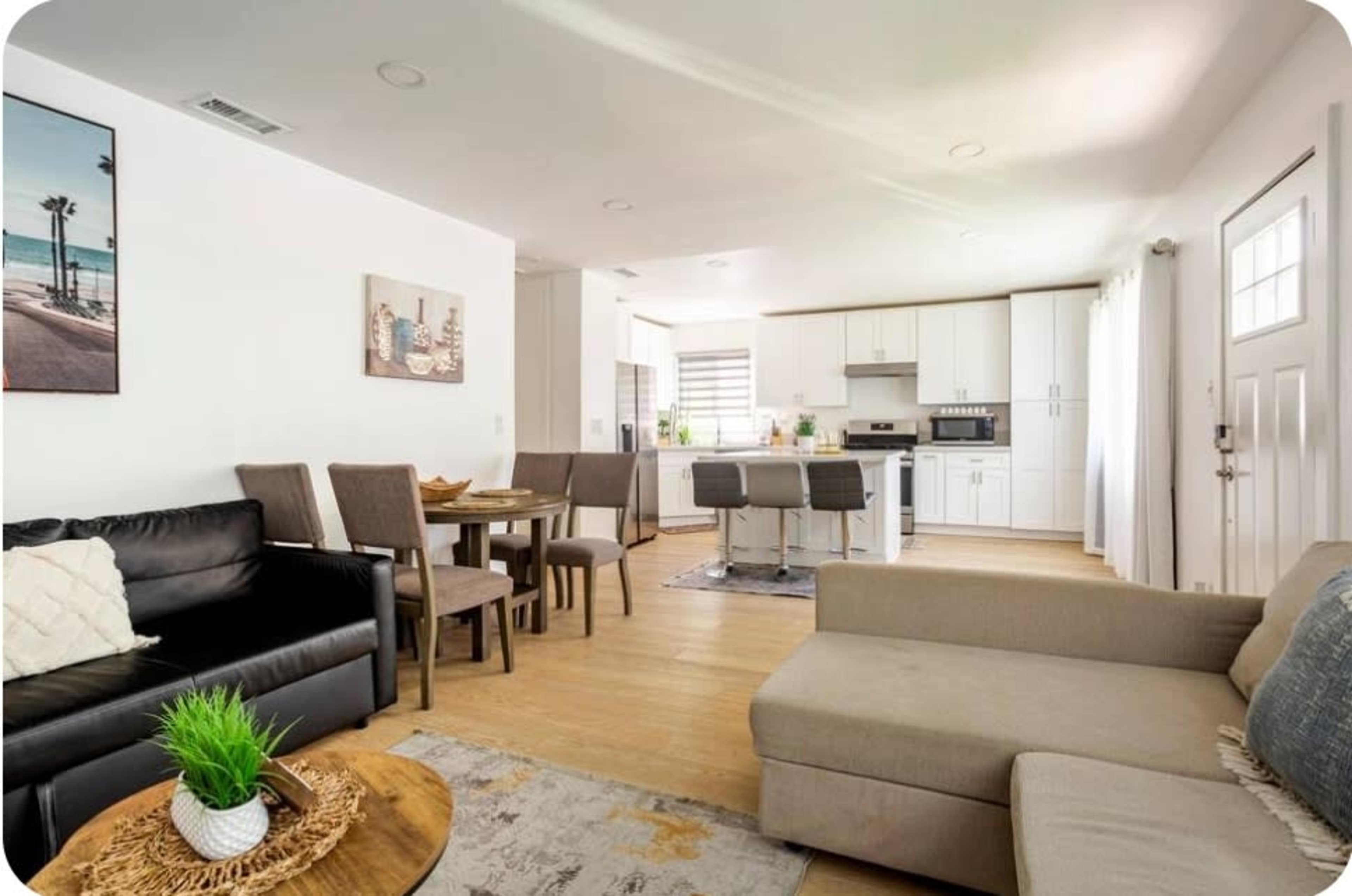 The image shows a modern living room and dining area with a sofa, chairs, a wooden table, and a kitchen visible in the background.