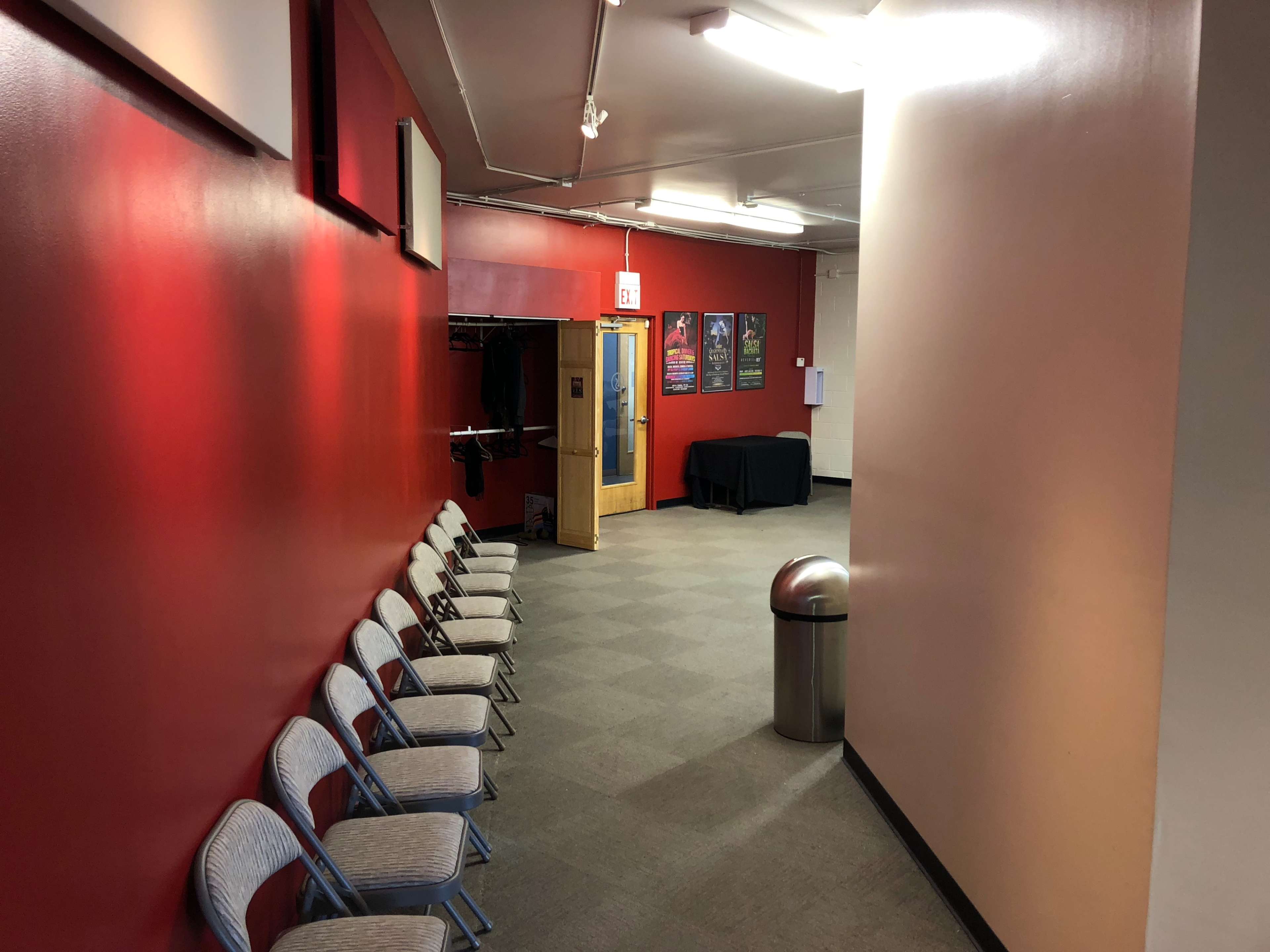 A long hallway with red walls, a row of folding chairs on one side, a trash can at the end, and a door leading to another room.