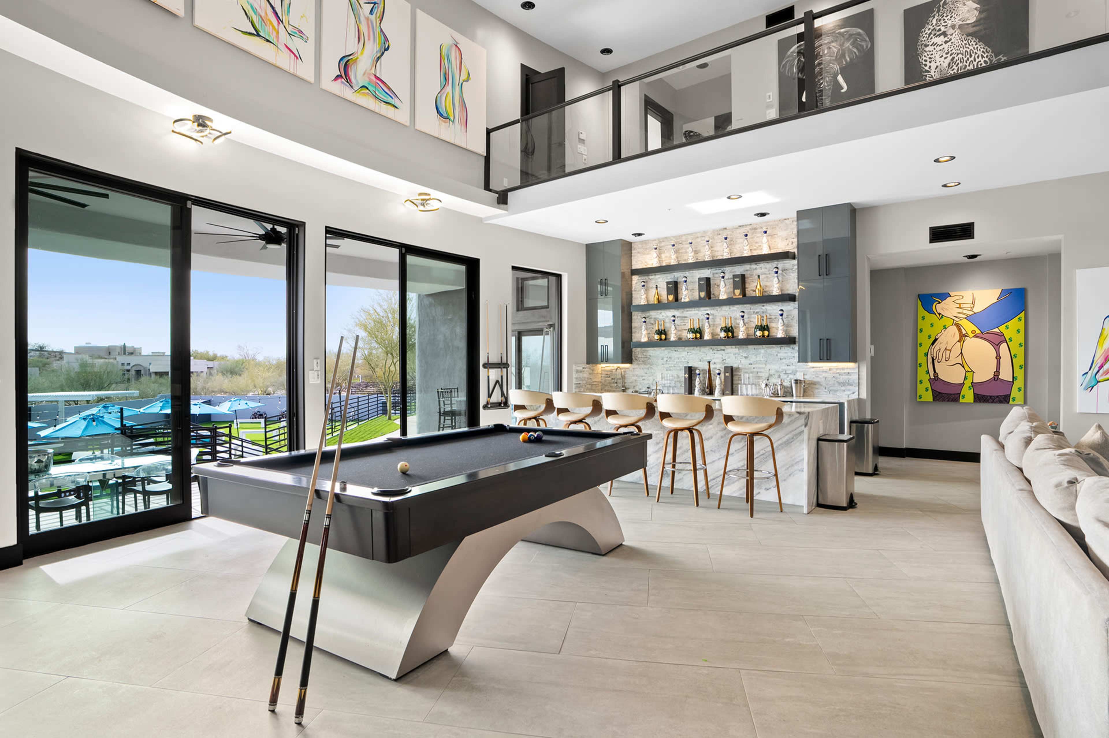 The image shows a modern interior featuring a pool table, a bar area with stools, large windows overlooking an outdoor space, and contemporary artwork on the walls.