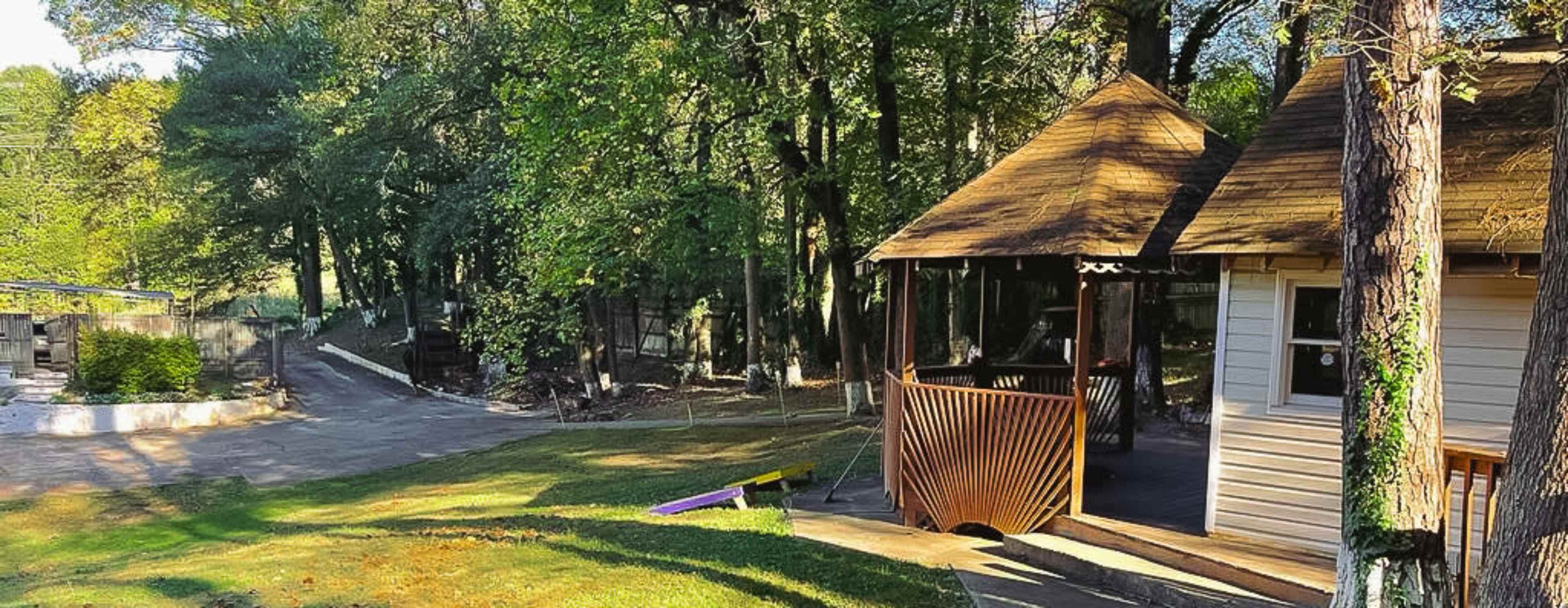 A quiet outdoor area with a wooden gazebo, a pathway, and a partially shaded landscape surrounded by trees.