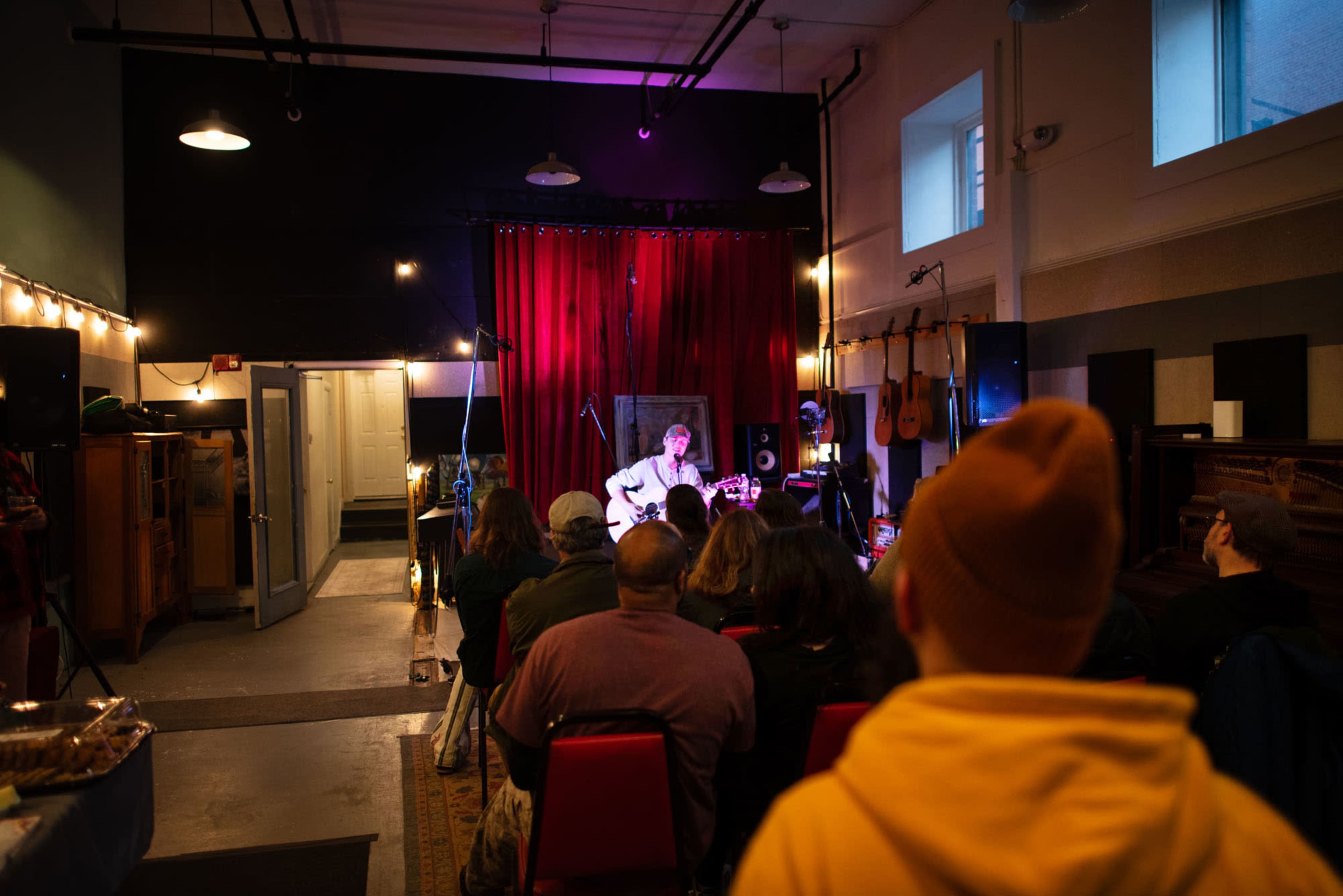 A musician plays guitar on stage in a dimly lit venue while an audience listens attentively.