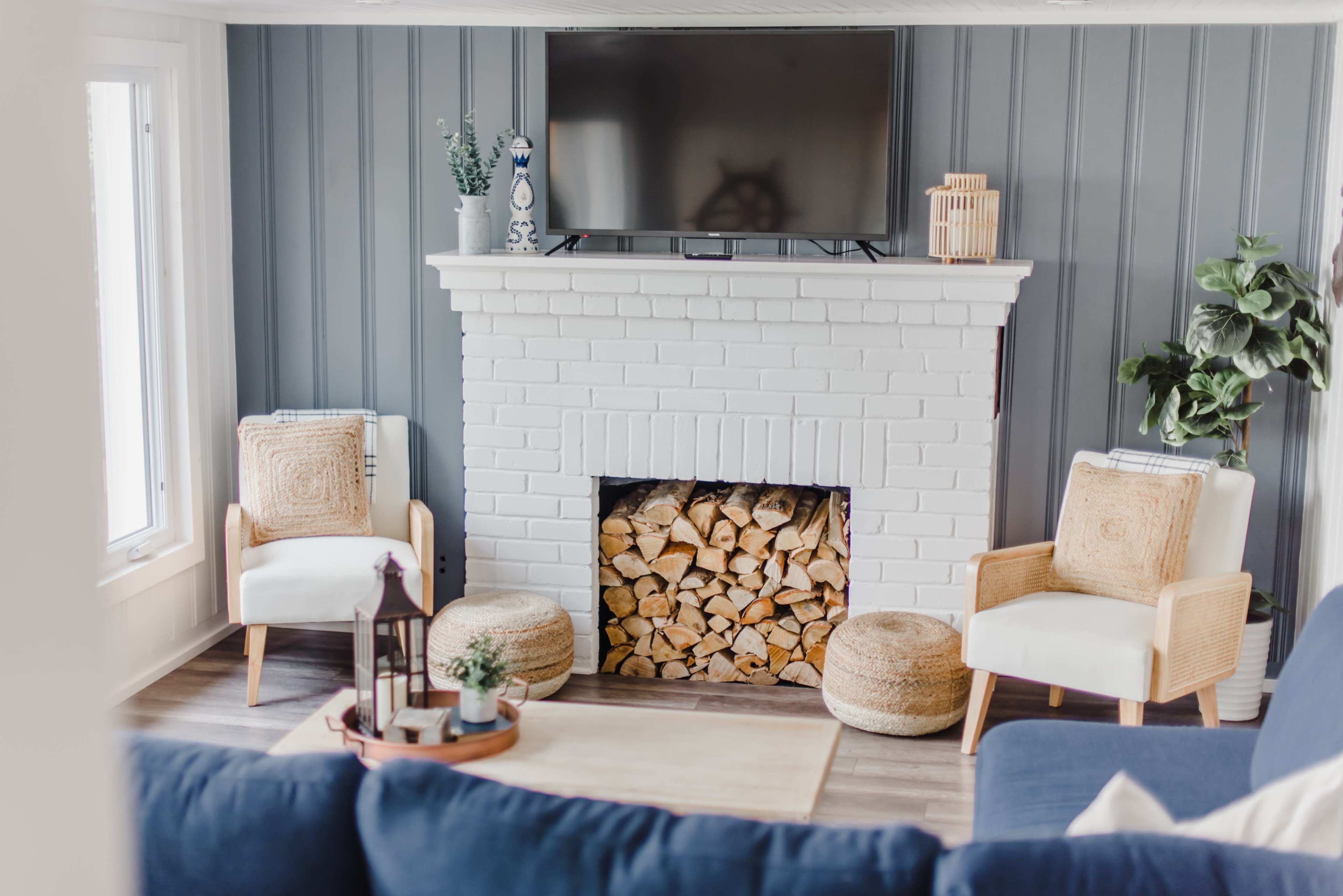 A living room with a white brick fireplace, a flat-screen TV mounted above it, and two light-colored chairs flanking a pile of stacked firewood.