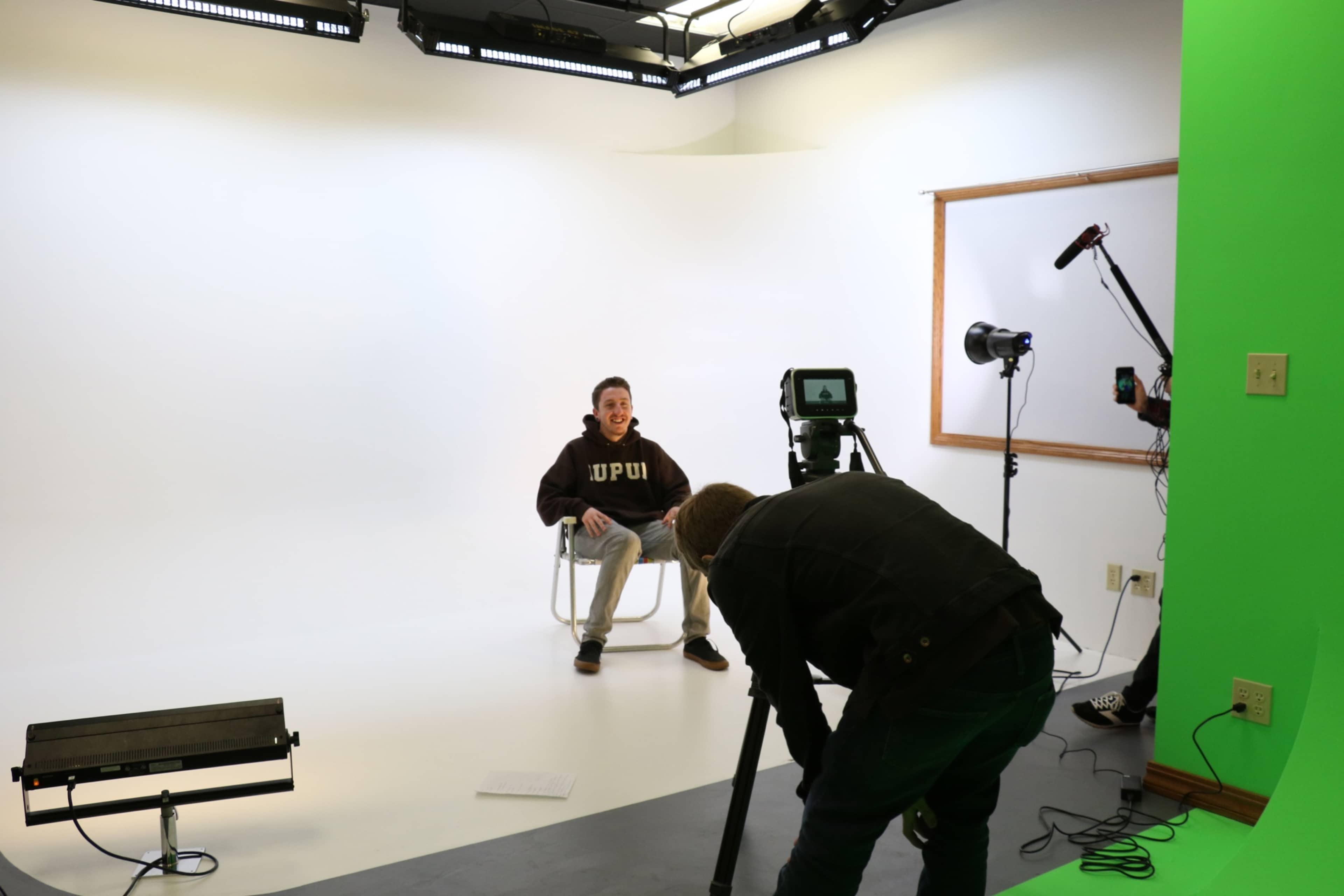 A seated individual in a hoodie is being filmed with a camera and lighting equipment in a studio environment.