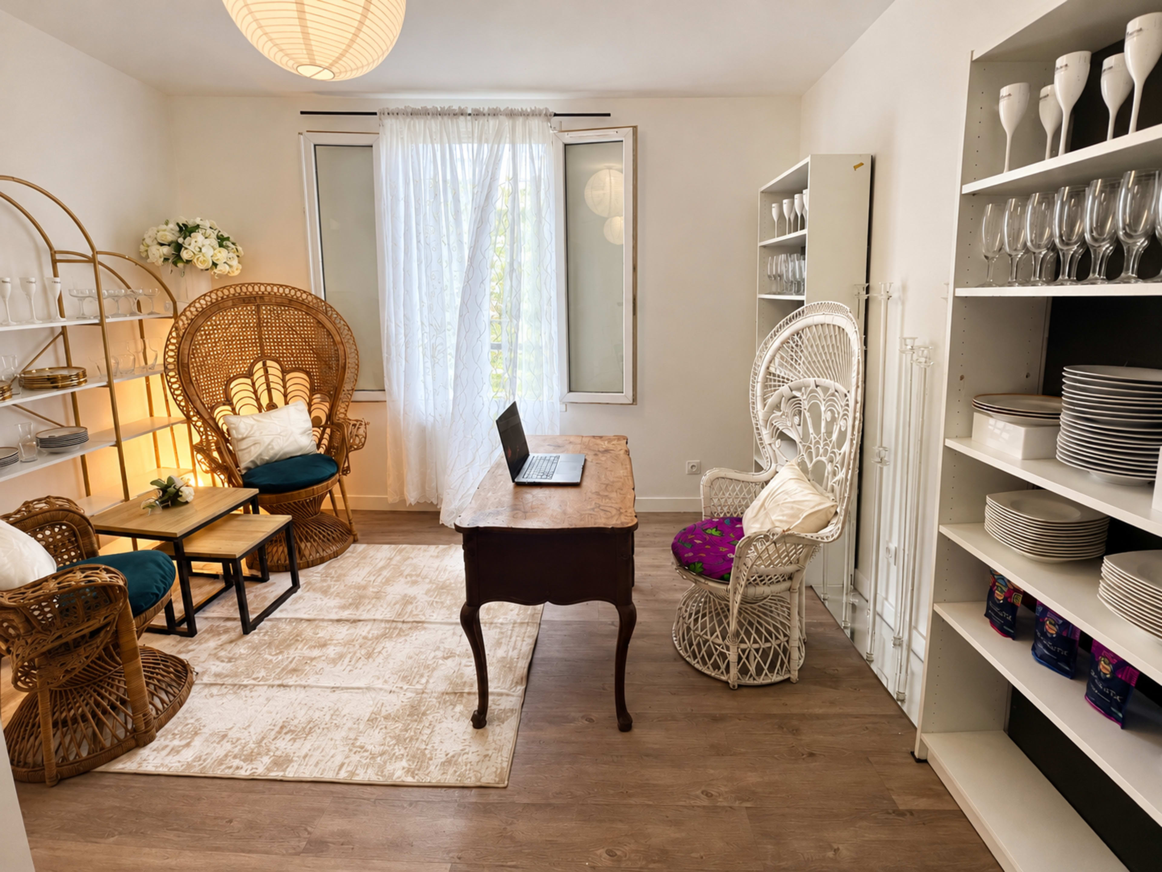 The room features a cozy seating area with two wicker chairs and a small table, a desk with a laptop, and shelving displaying glassware and dinnerware.