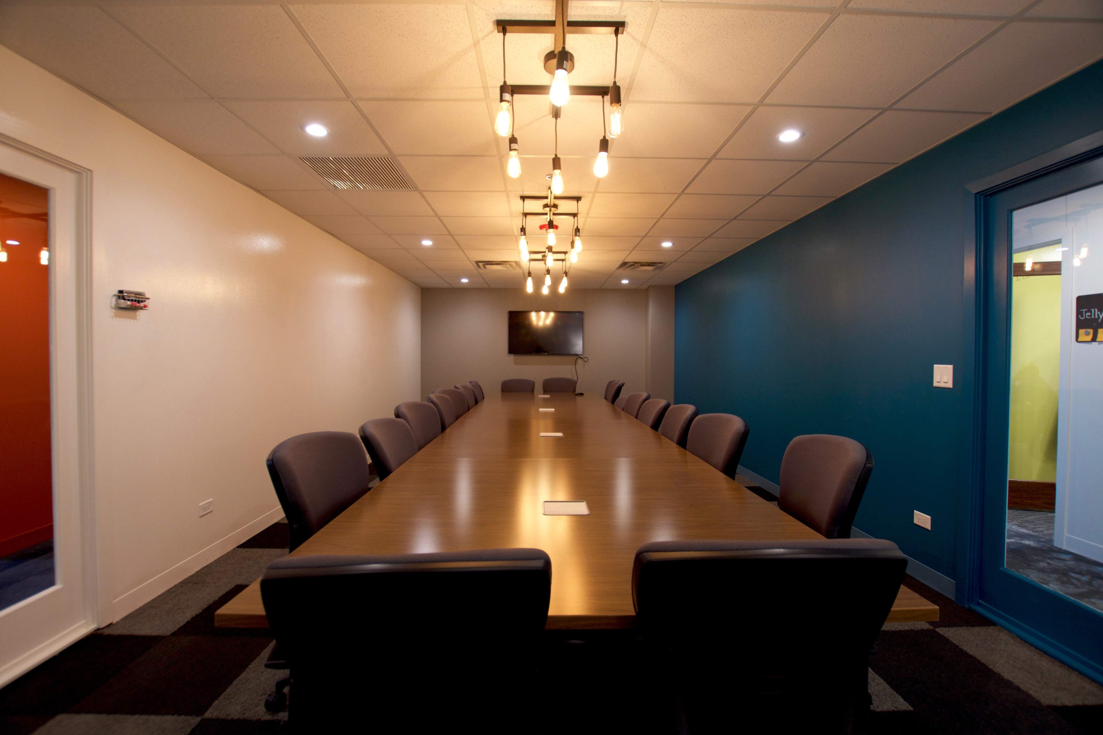 A long conference table with dark chairs is positioned in a well-lit meeting room featuring blue and white walls and a television mounted on one wall.