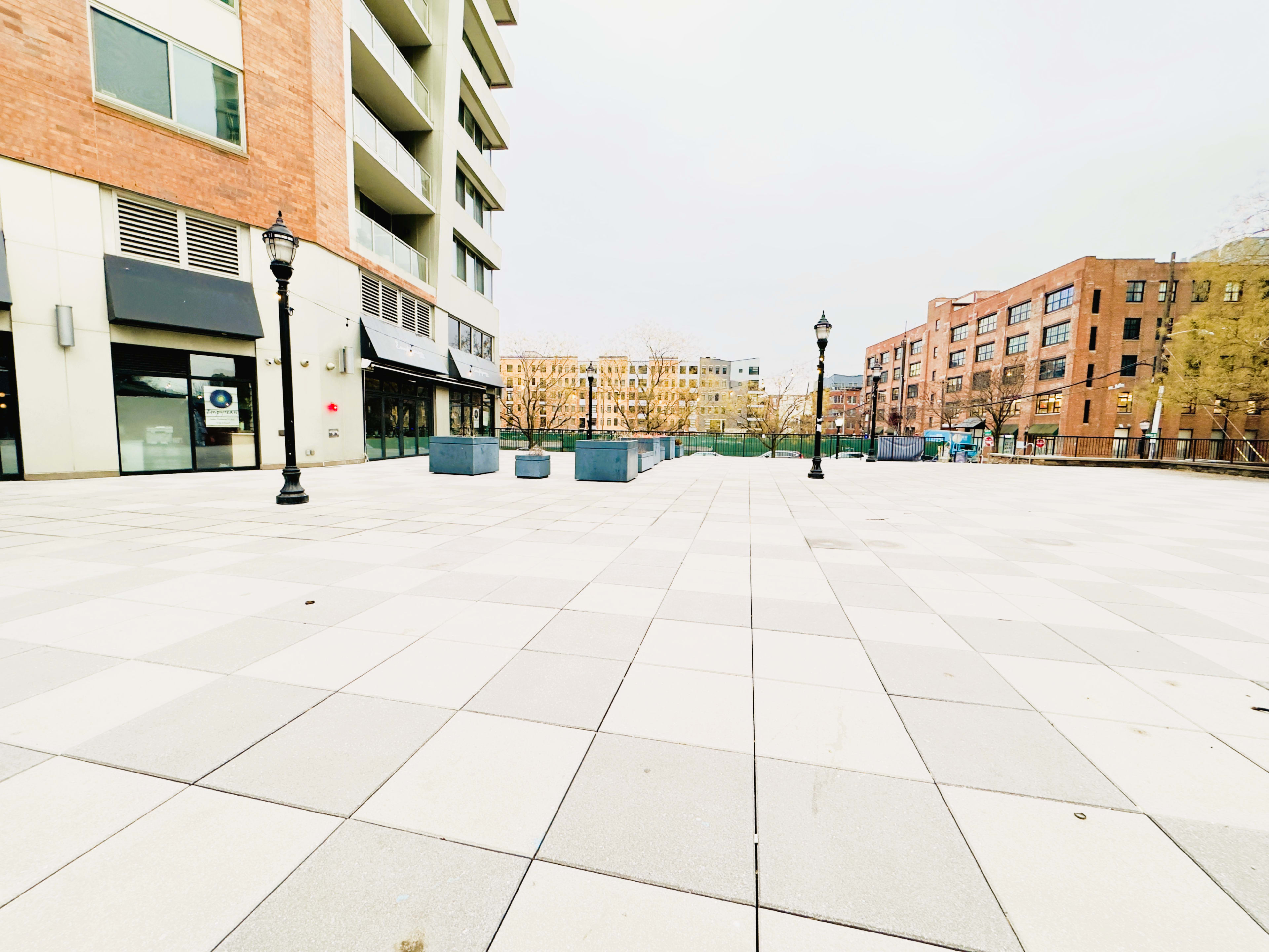 The image shows a spacious outdoor plaza with large gray paving stones, surrounded by buildings and lampposts.