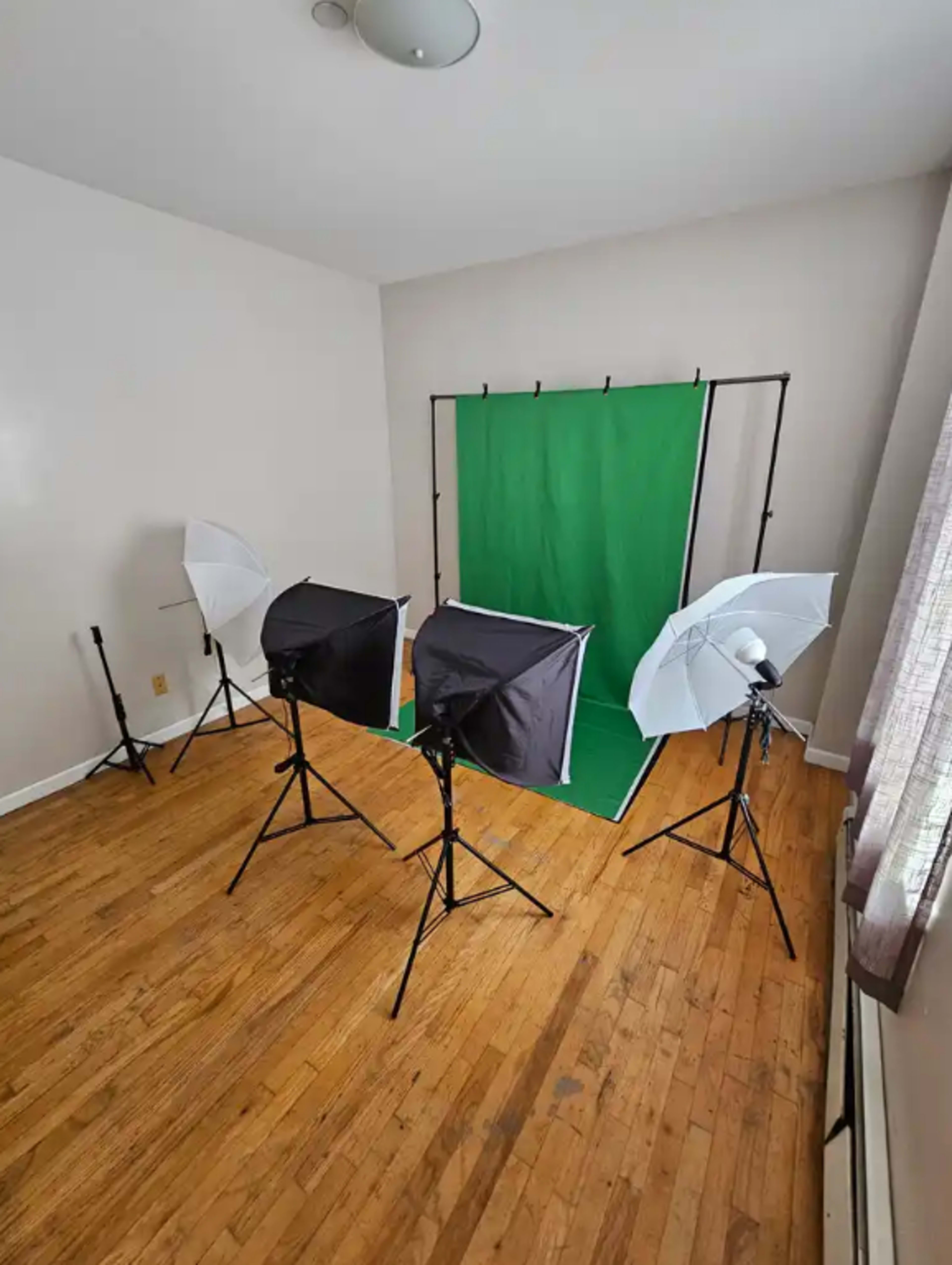 The image shows a studio setup with a green backdrop surrounded by four lighting umbrellas on stands, positioned in a room with wooden floors.