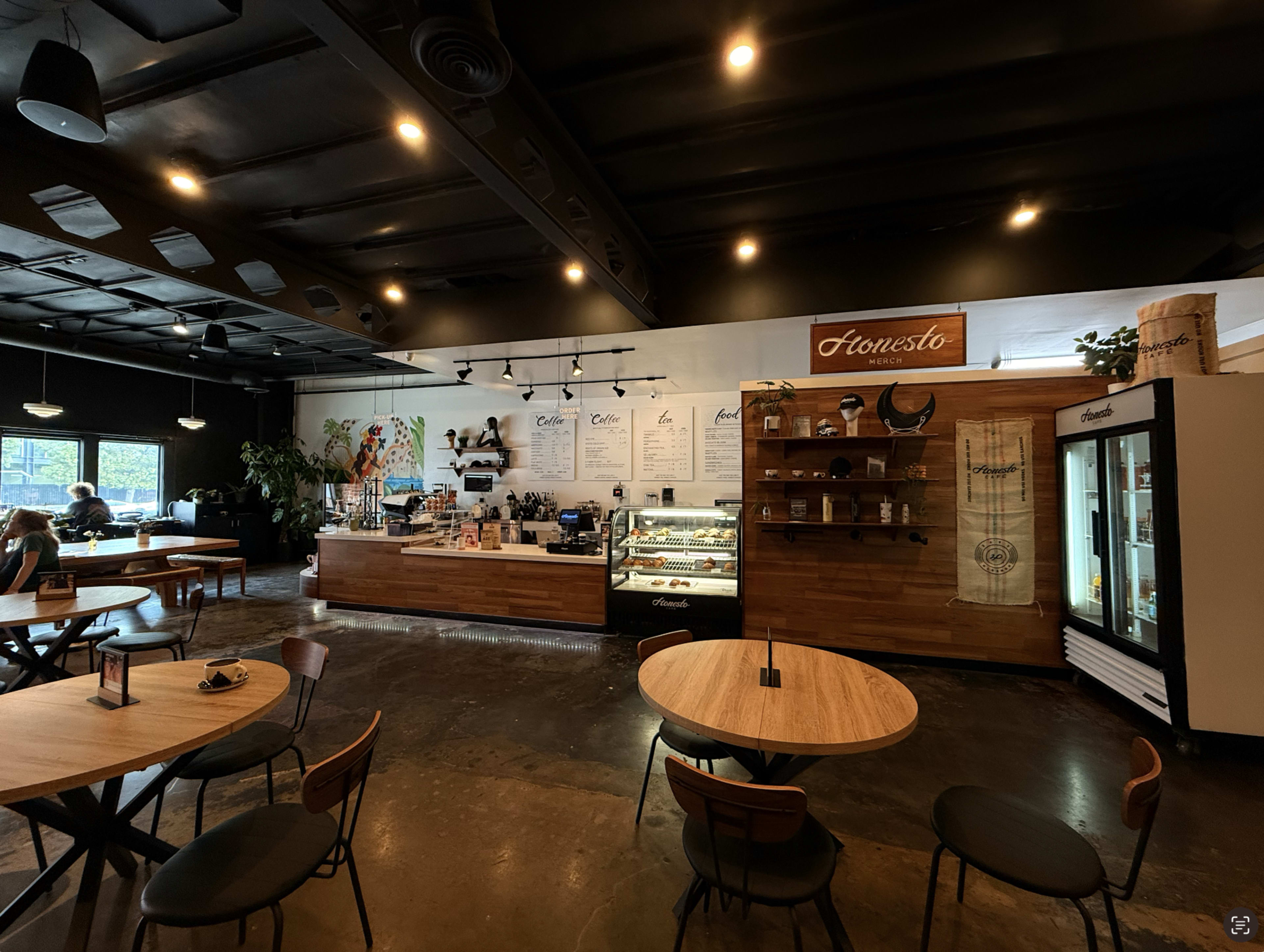 The image depicts a modern café interior featuring wooden furniture, a display counter with pastries, and menu boards on the wall.
