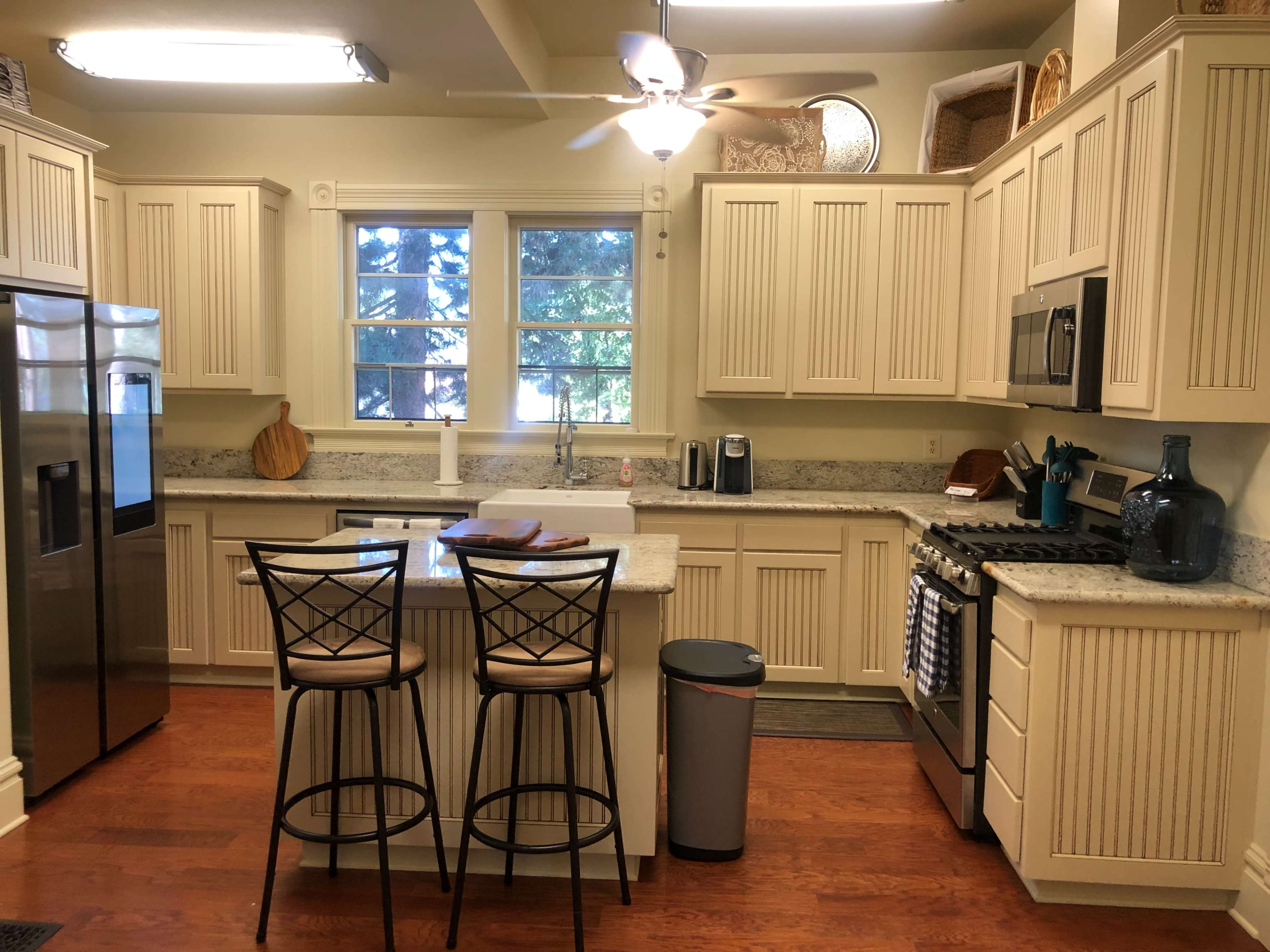 A modern kitchen features light-colored cabinetry, a granite countertop island with two bar stools, and stainless steel appliances.