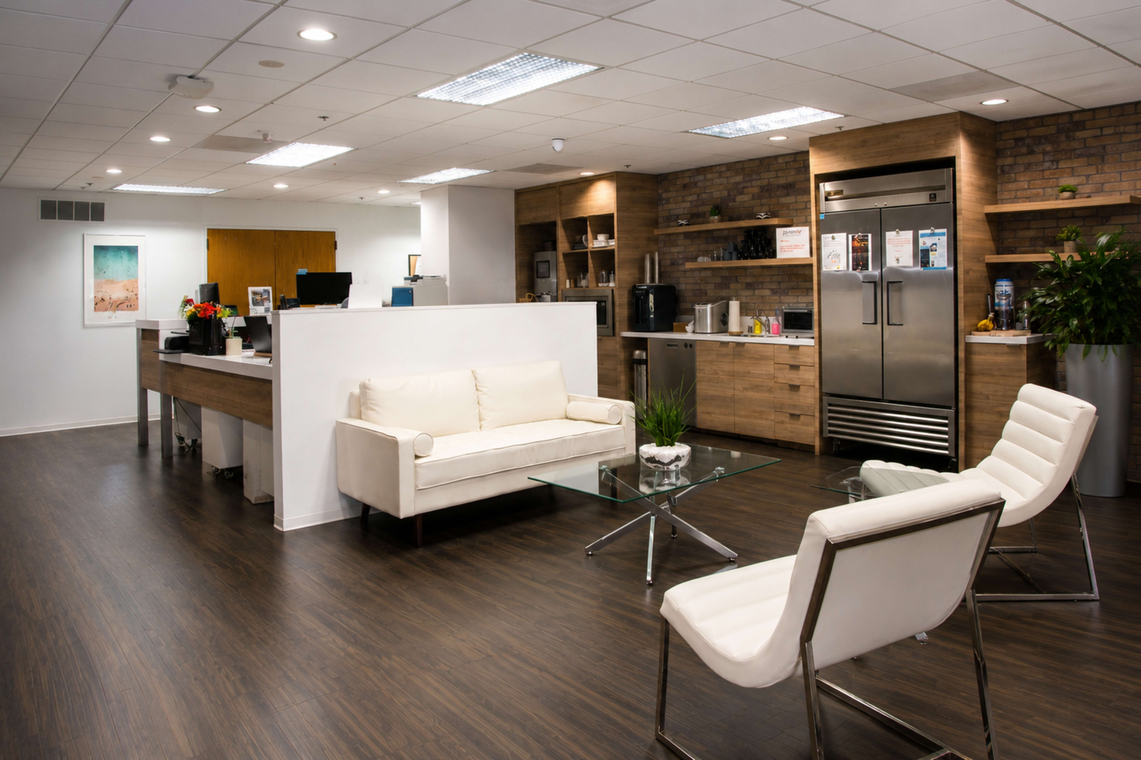 The image shows a modern office break room with a white sofa, glass coffee table, and a kitchenette featuring wood accents and stainless steel appliances.