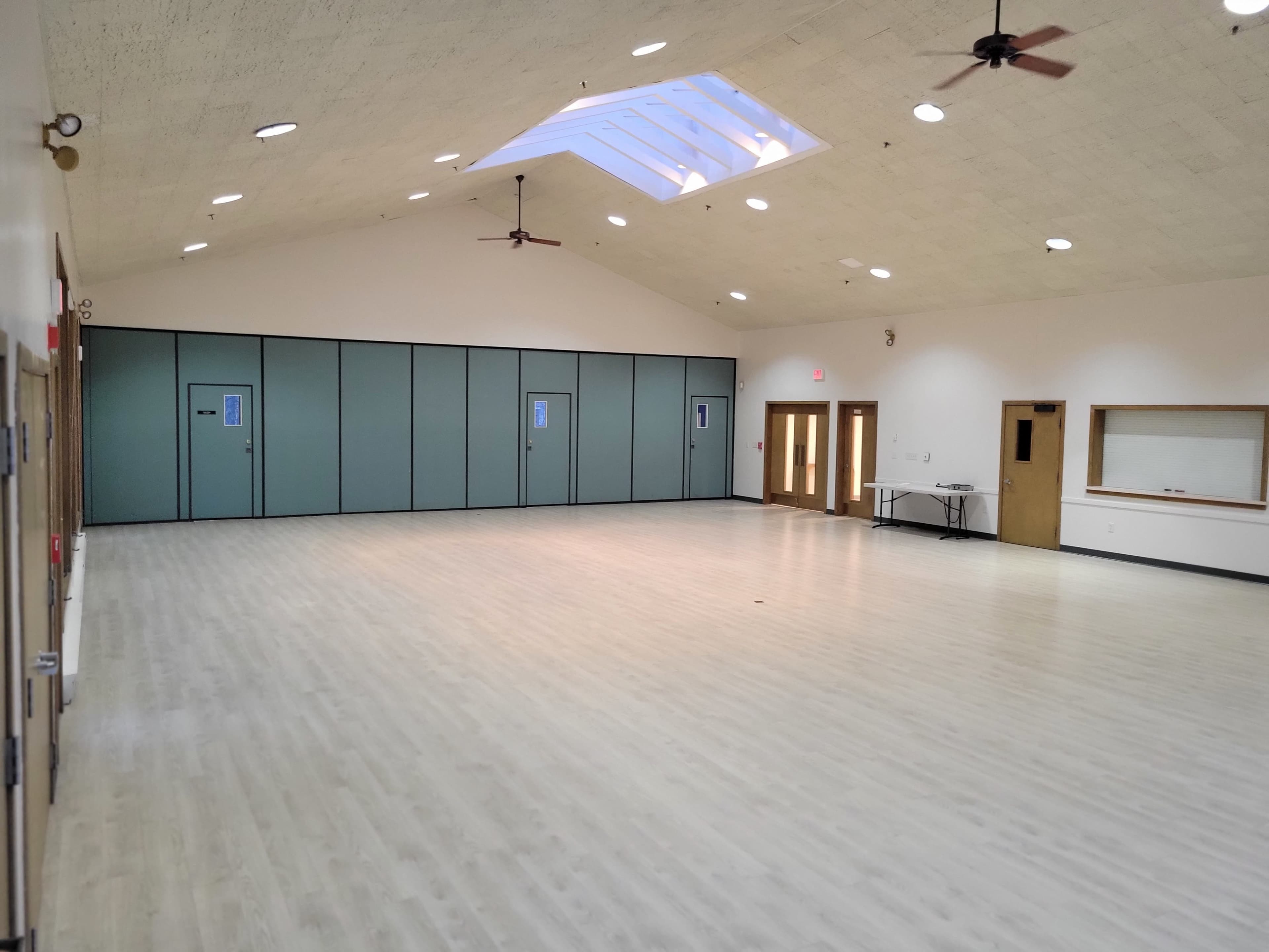 The image shows a spacious indoor room with a high ceiling, large windows, and a wood floor, featuring two sets of double doors and a small table against the wall.