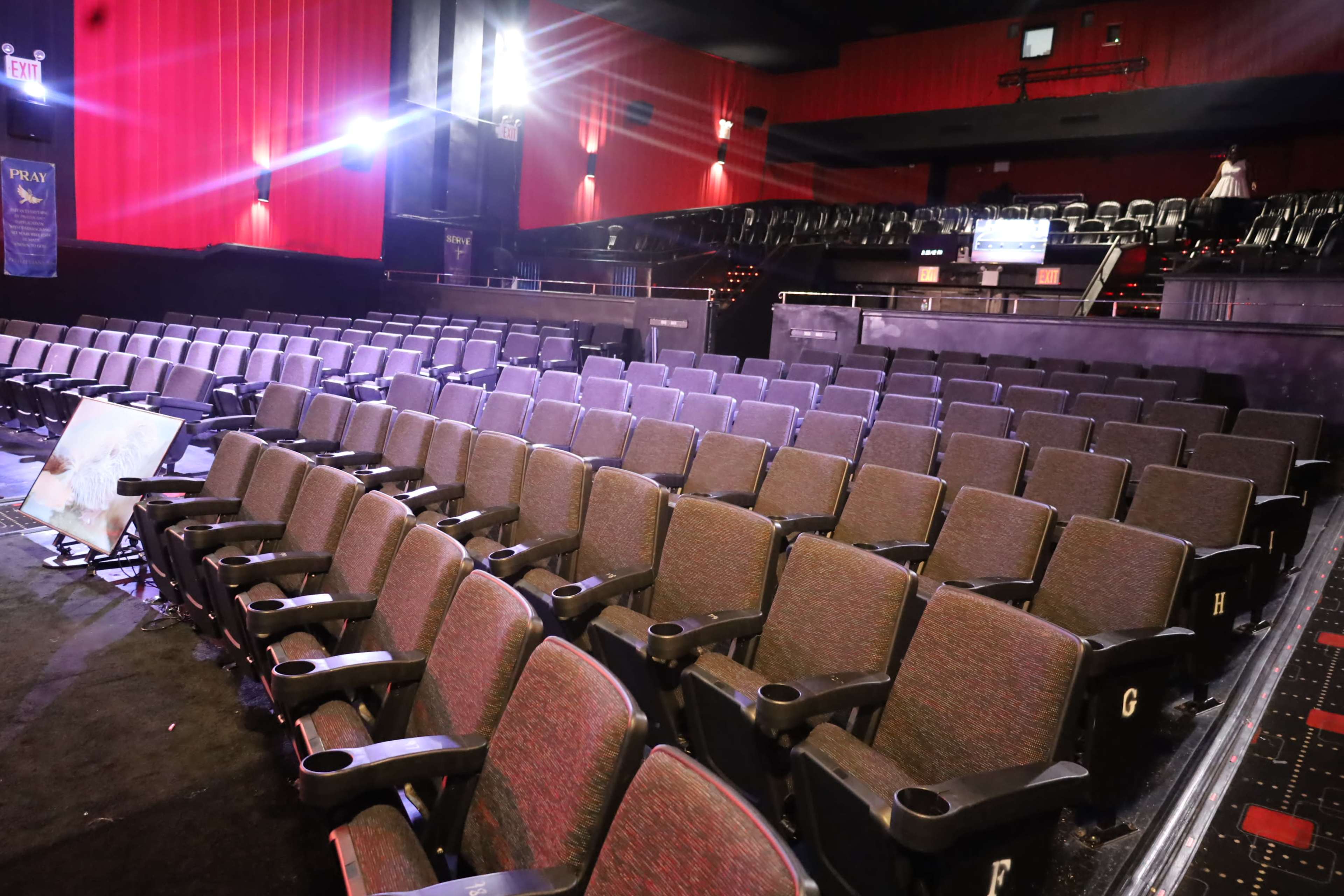 The image shows a nearly empty movie theater auditorium with rows of red and black seats, illuminated by overhead lights.